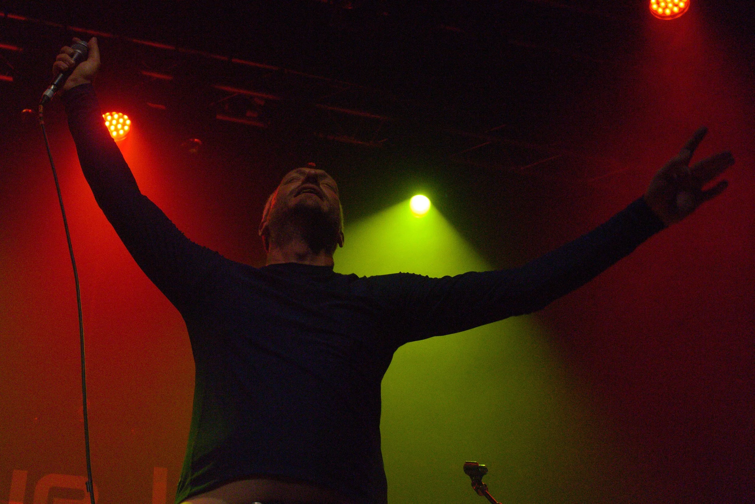 Male singer performing on stage with microphone in his right hand, arms raised, wearing a black long sleeve shirt, in front of red and green stage lighting.