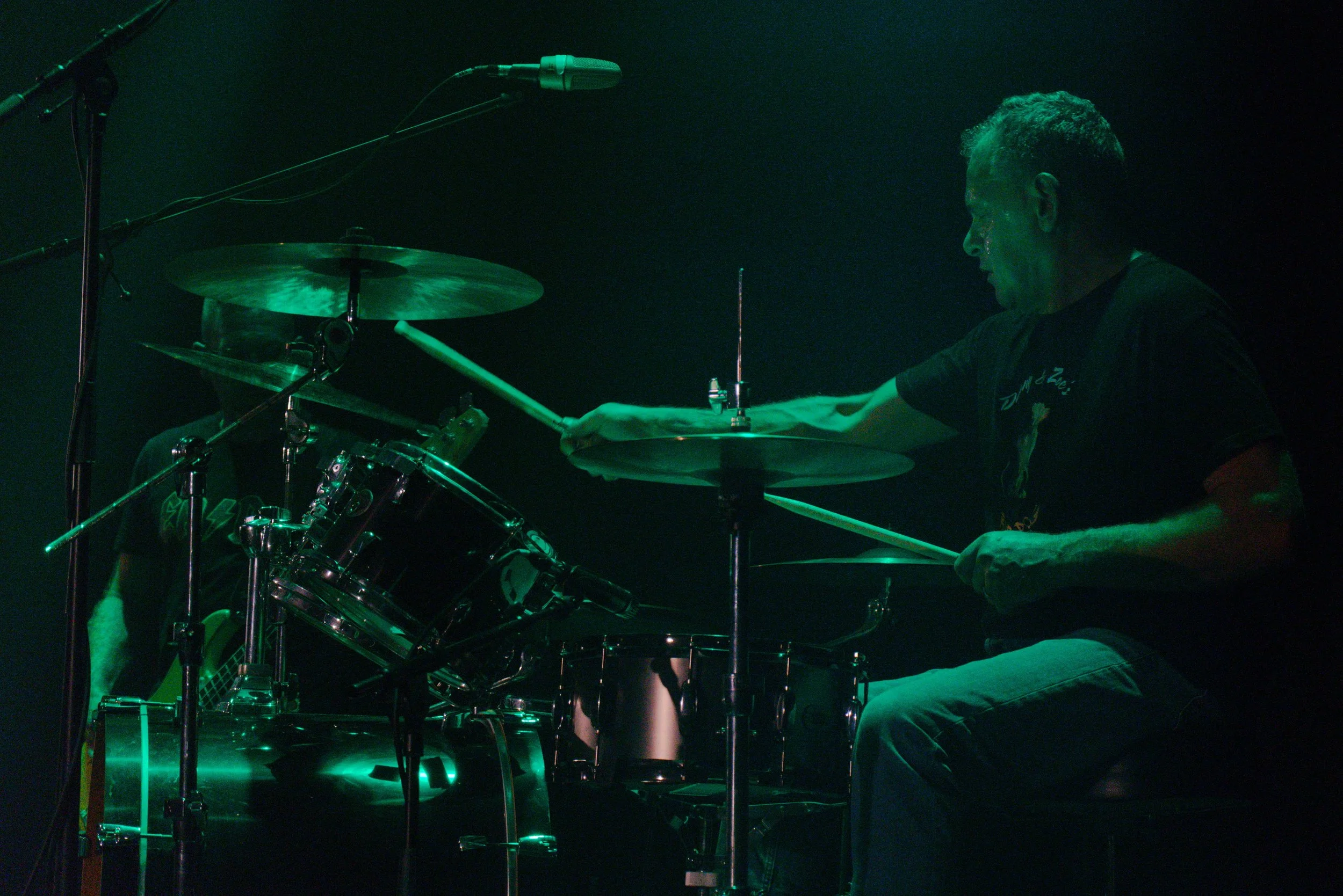 A drummer playing on stage with dark background, green stage lighting, drum set, and microphone.