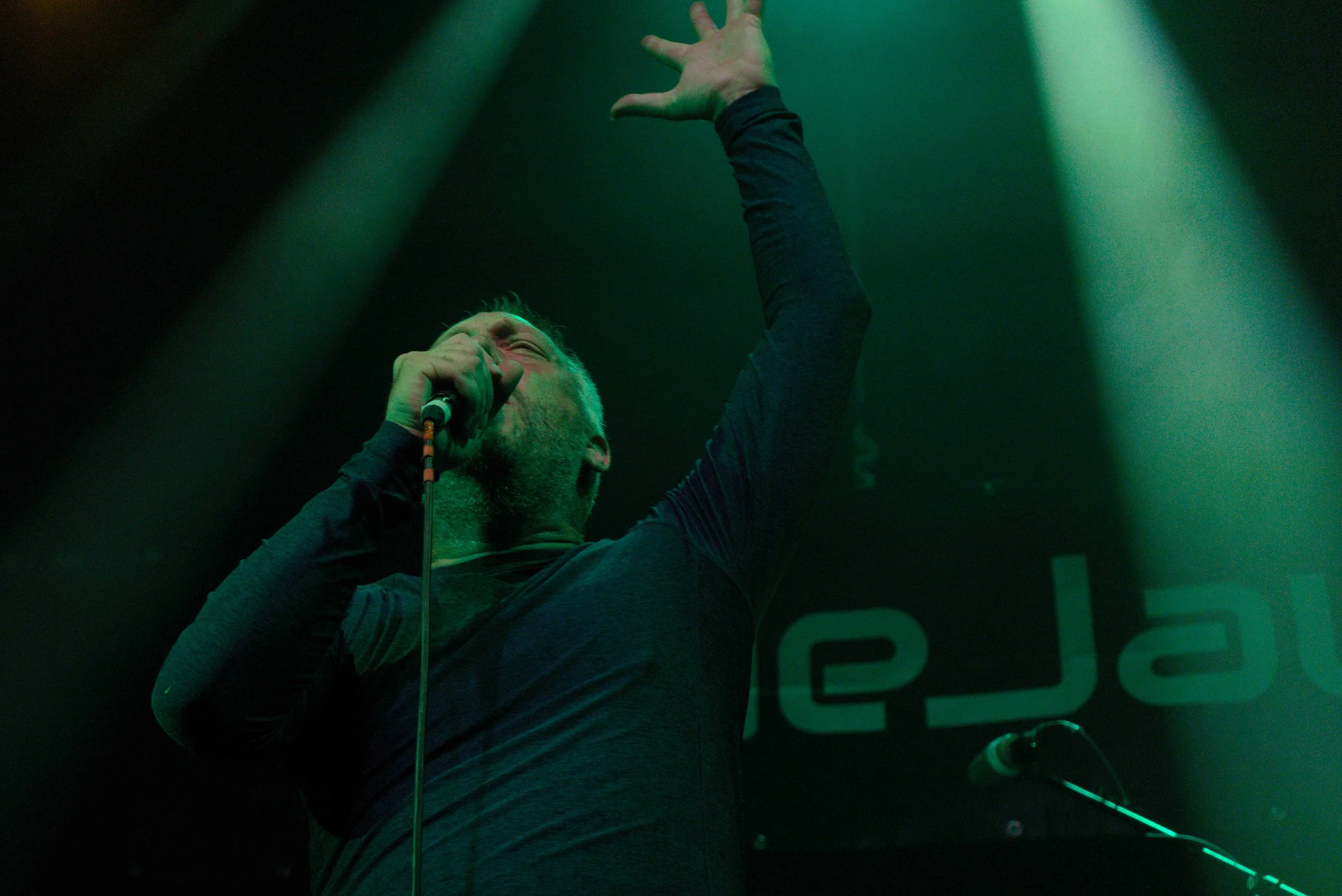 Male singer with grey hair singing into a microphone on stage, with one arm raised, under green stage lighting.