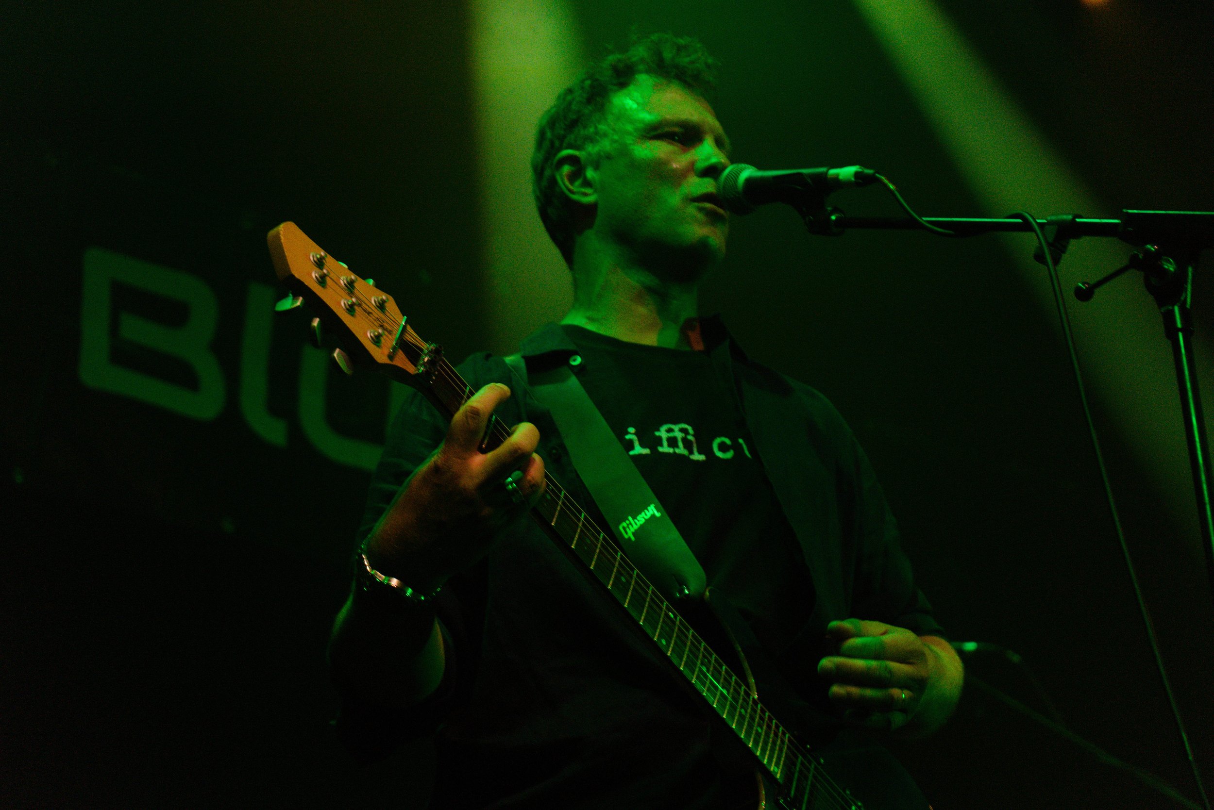 Man singing into a microphone while playing an electric guitar on stage with green lighting