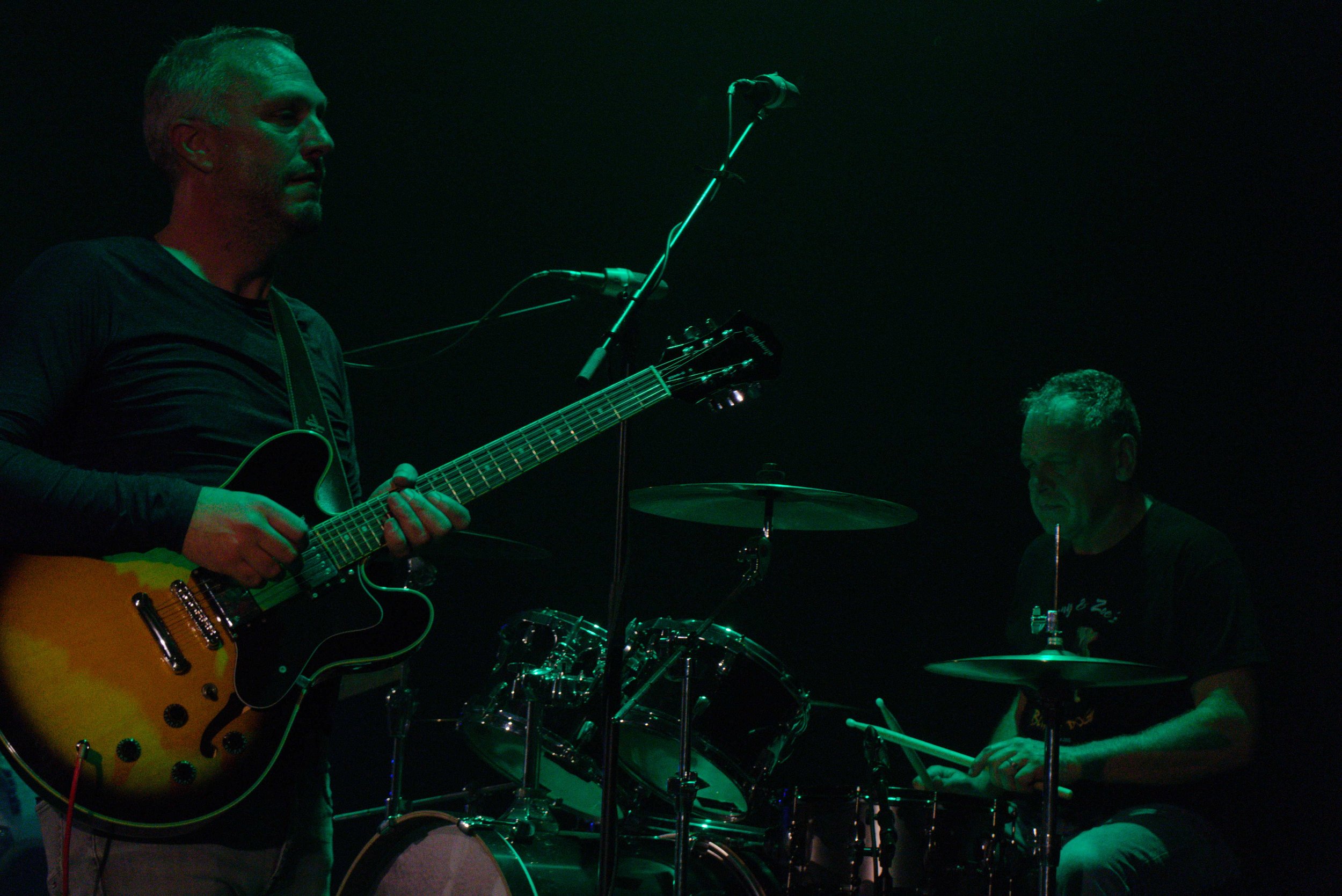 Two musicians performing on stage with dark background are playing instruments, one with an acoustic guitar and the other with drums.