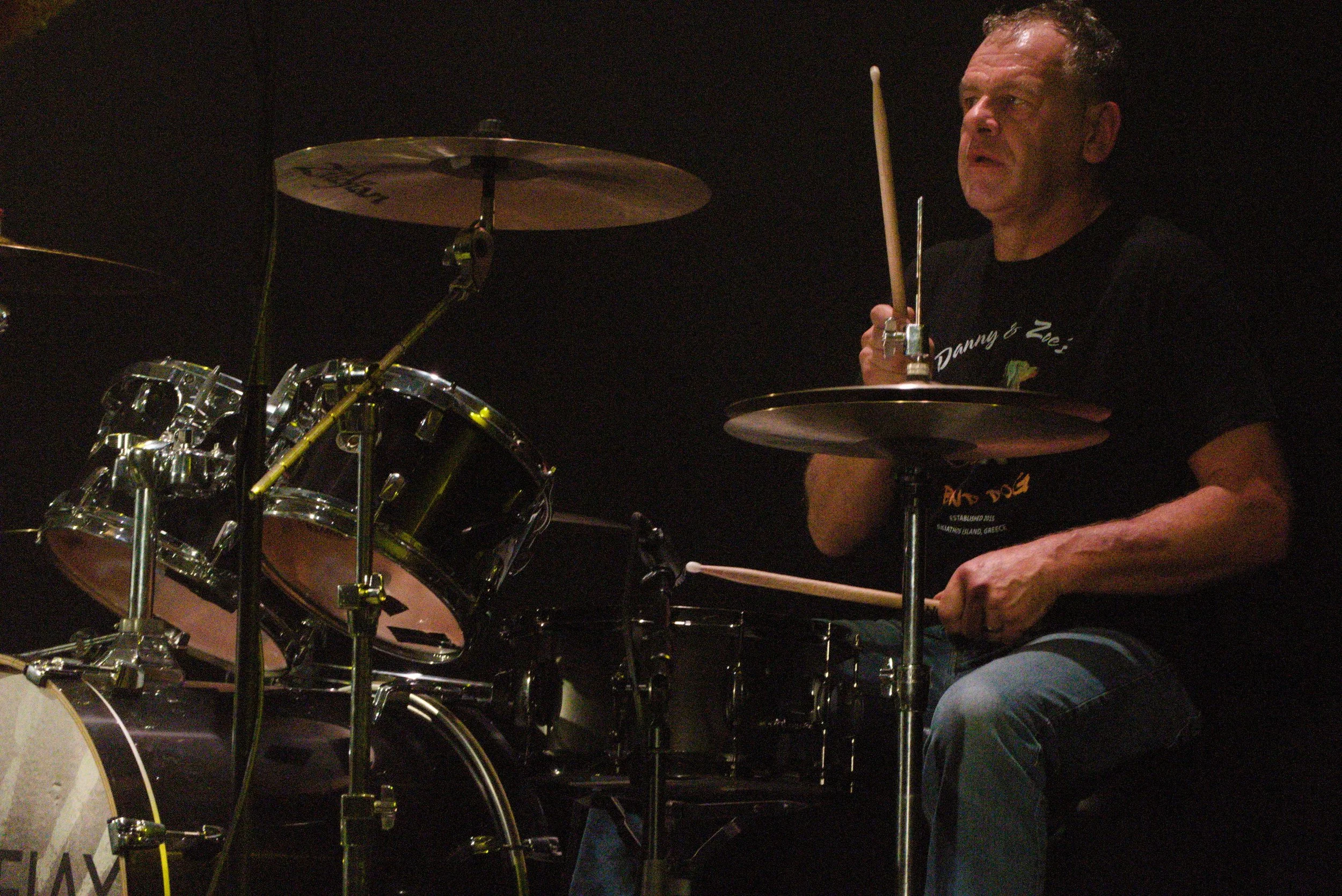 A man playing the drums on stage, holding drumsticks, with a drum set in front of him, wearing a black t-shirt with text, with a dark background.