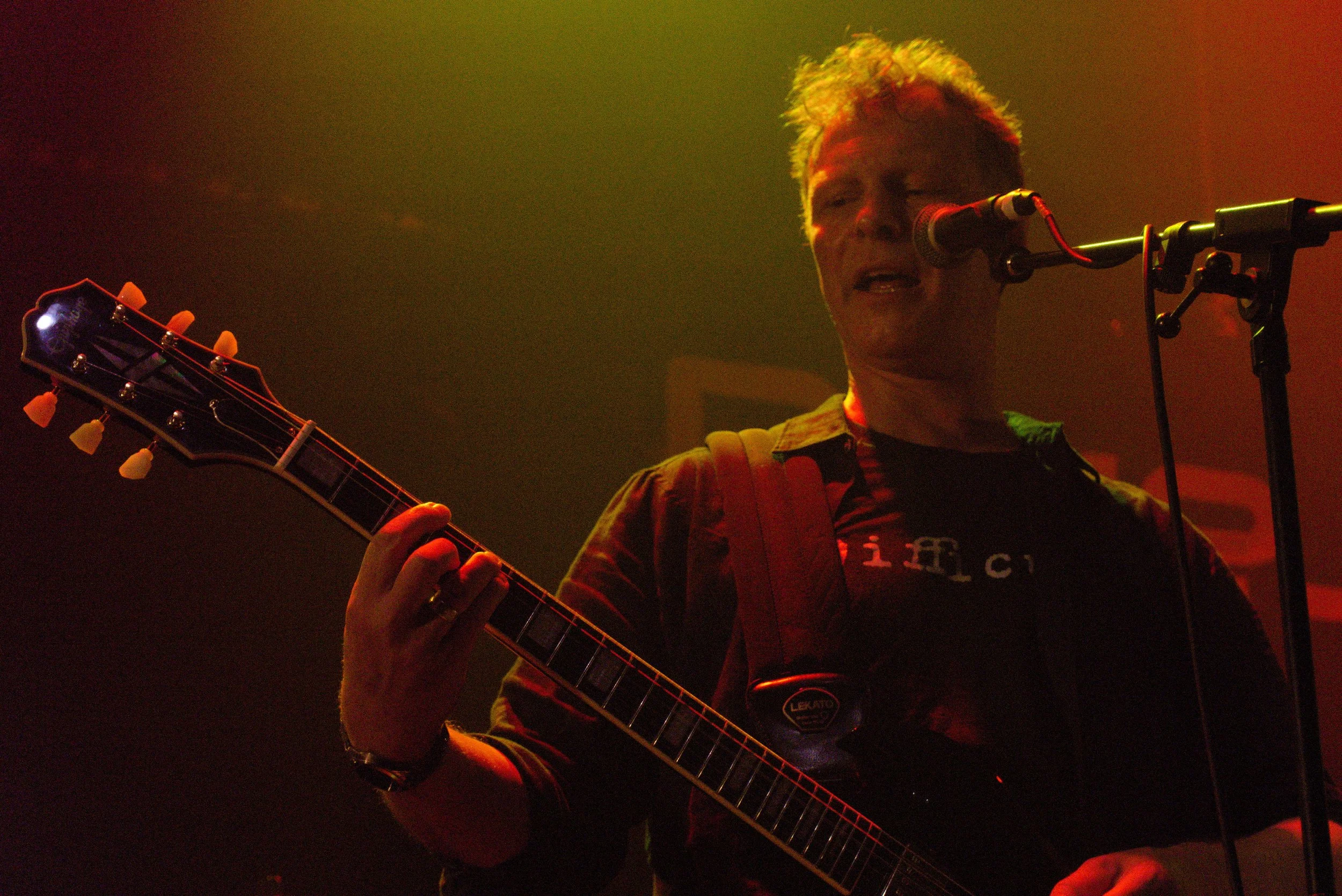 A person with blonde hair singing into a microphone while playing an acoustic guitar on stage, with red and yellow stage lighting.