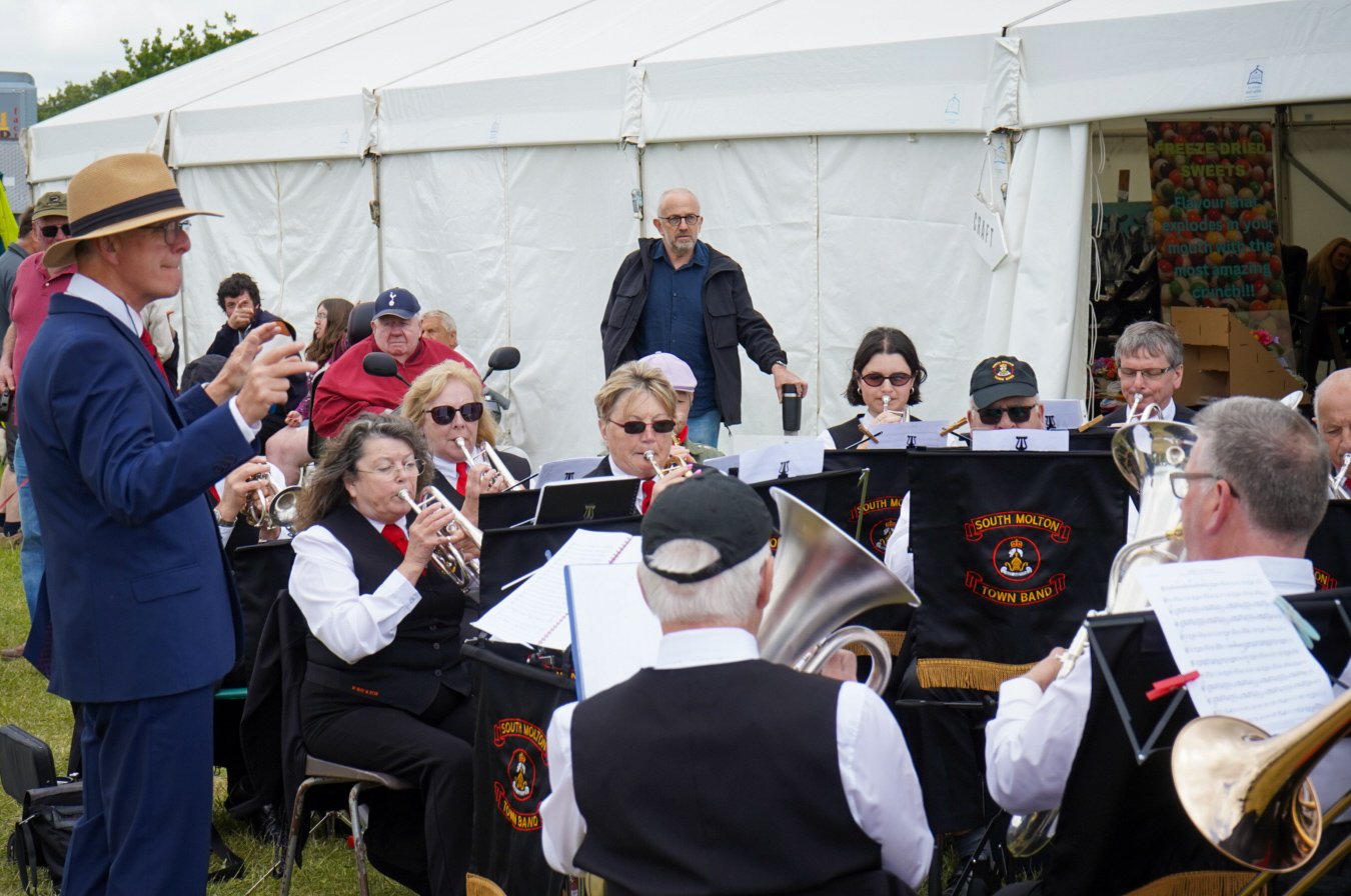 A musical band performing outdoors at a community event, with a conductor in a blue suit and straw hat leading them.