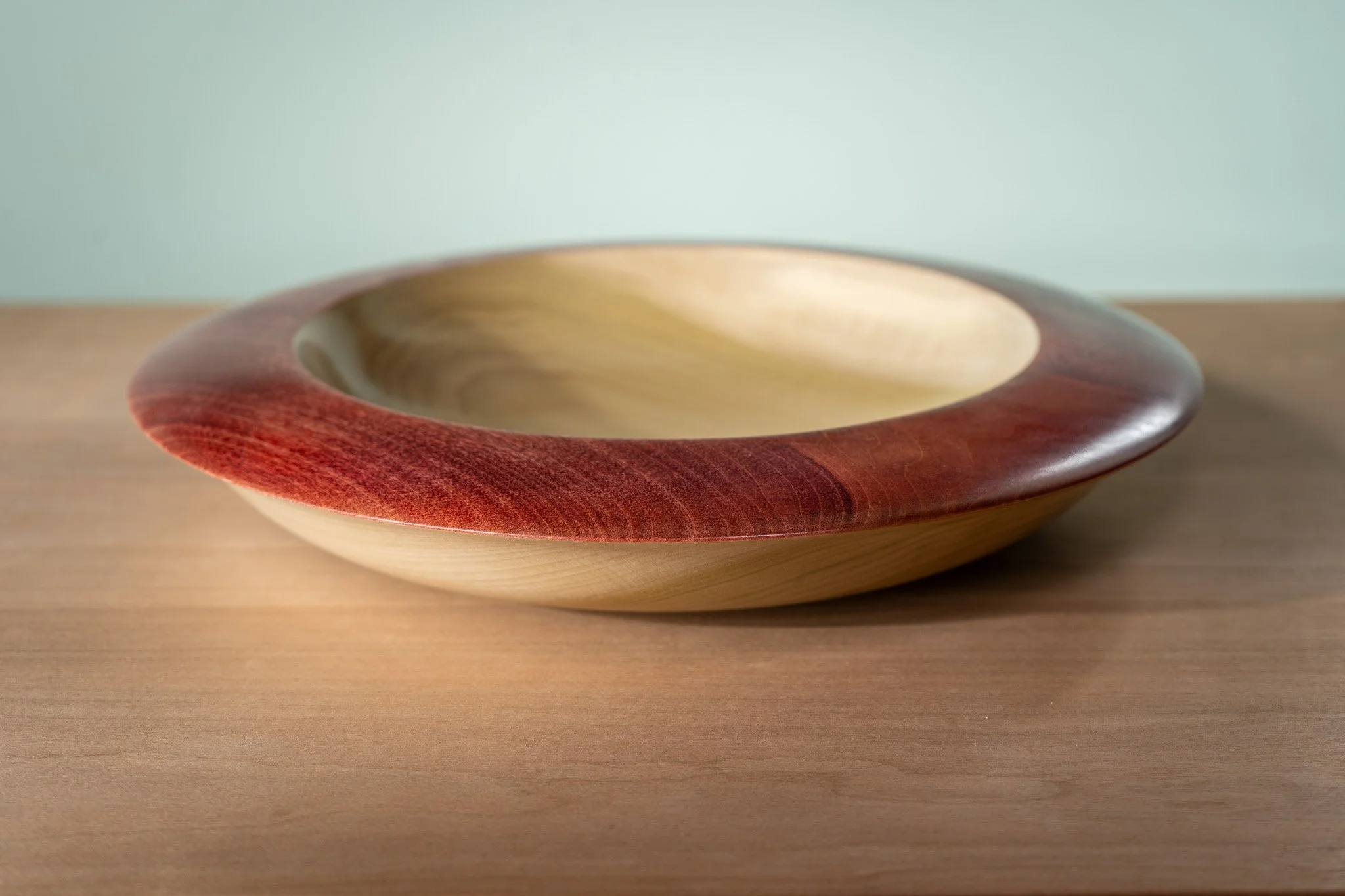 A wooden bowl with a reddish-brown exterior and a light-colored interior, placed on a wooden surface with a light green background.