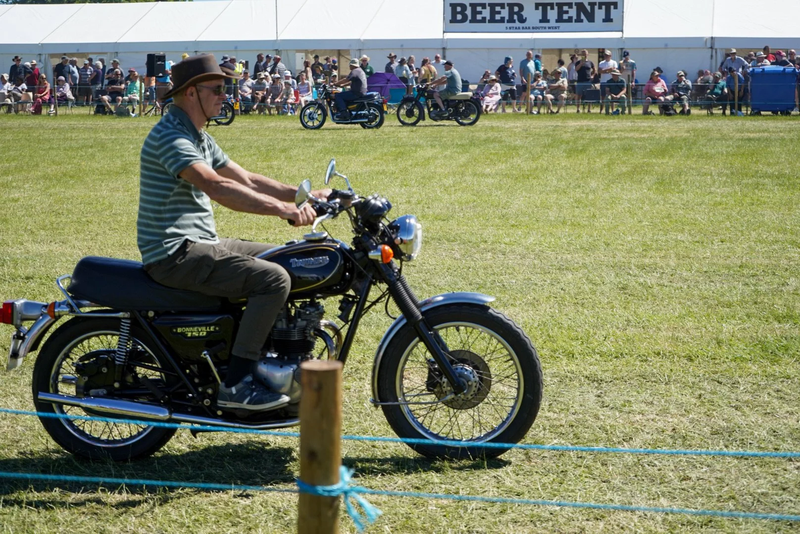 A man wearing a striped shirt, brown hat, and sunglasses riding a black vintage Triumph Bonneville 750 motorcycle across a grassy field at an outdoor event with a large crowd seated in front of a white tent that has a sign reading 'BEER TENT'.