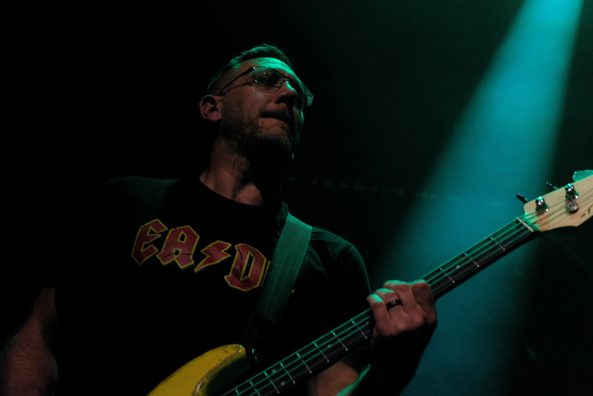 A man playing an electric bass guitar on stage, illuminated by green stage lights, wearing glasses and a black T-shirt with yellow and red lettering.