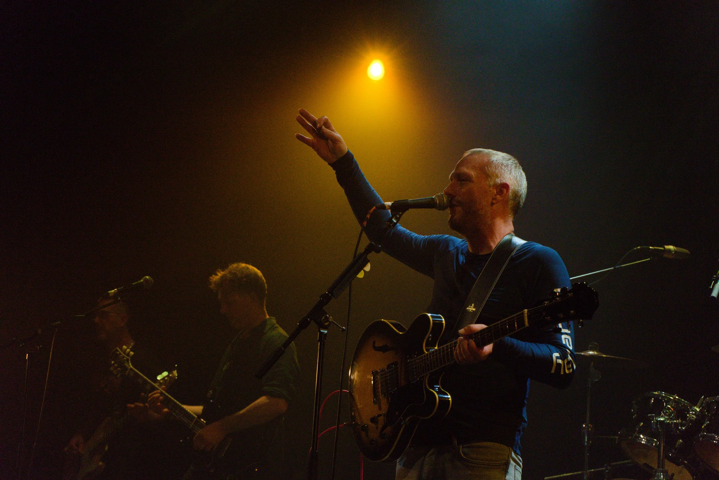 A man with short gray hair singing into a microphone while playing an acoustic guitar on stage, raising his hand with a peace sign gesture, with stage lighting and other musicians visible in the background.