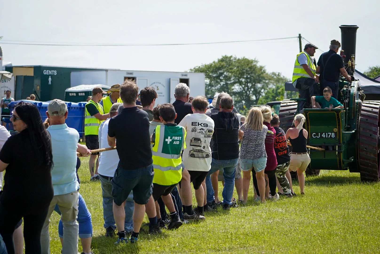 People participating in a tug-of-war game at an outdoor event, with one team pulling on a rope against a vintage tractor, and spectators observing. There are tents, portable toilets, and a group of people in the background.