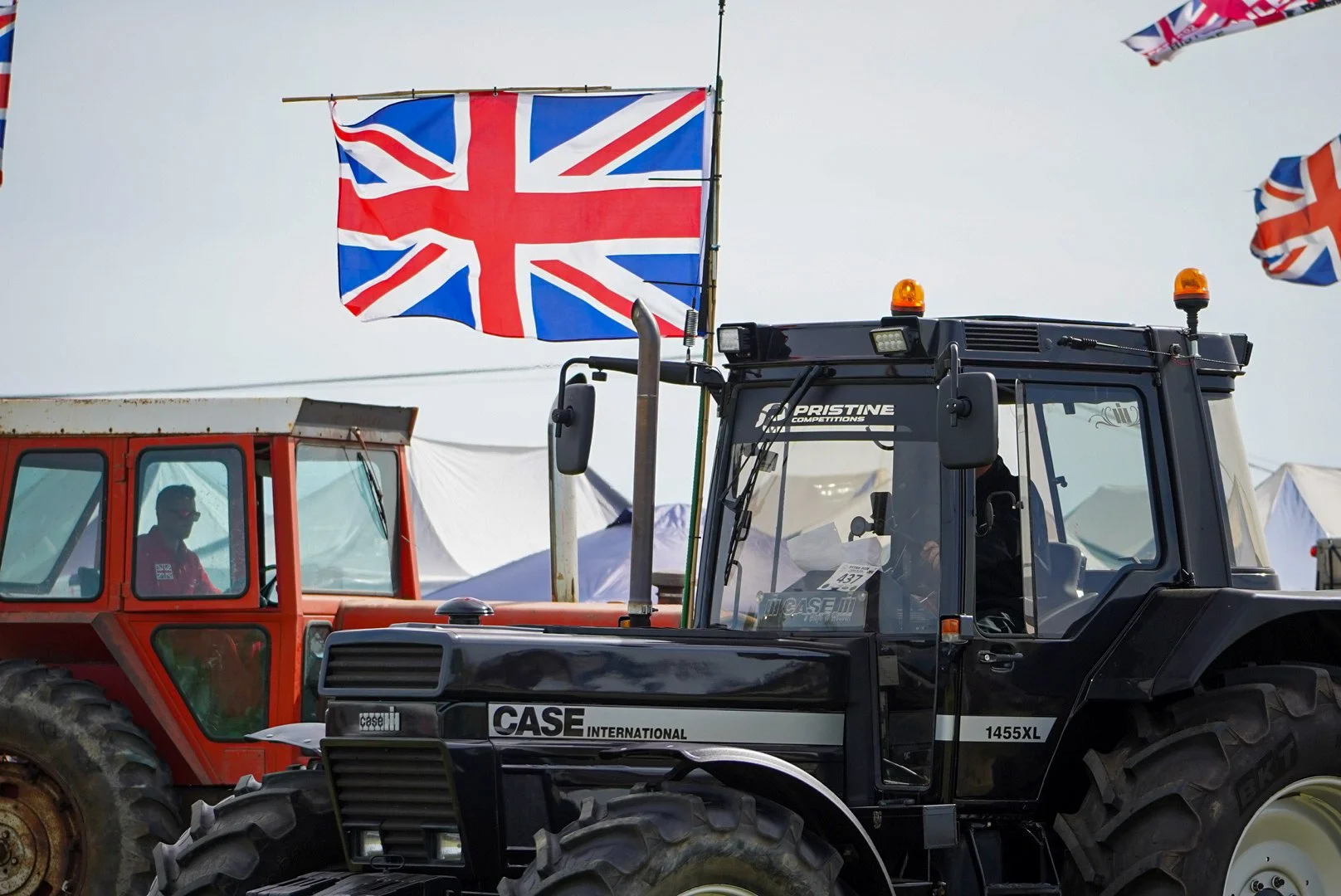 Close-up of a black CASE tractor with orange warning lights, with a red tractor and a British flag flying in the background during a farming event.