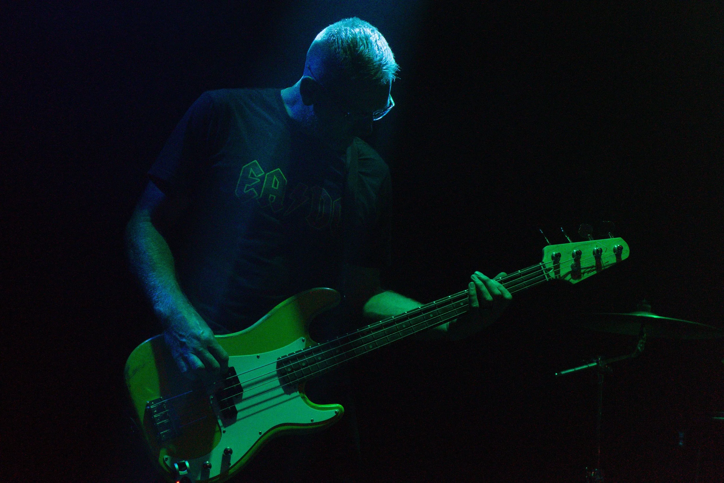 A person with short blonde hair playing a yellow electric bass guitar in a dark setting with blue and green lighting.