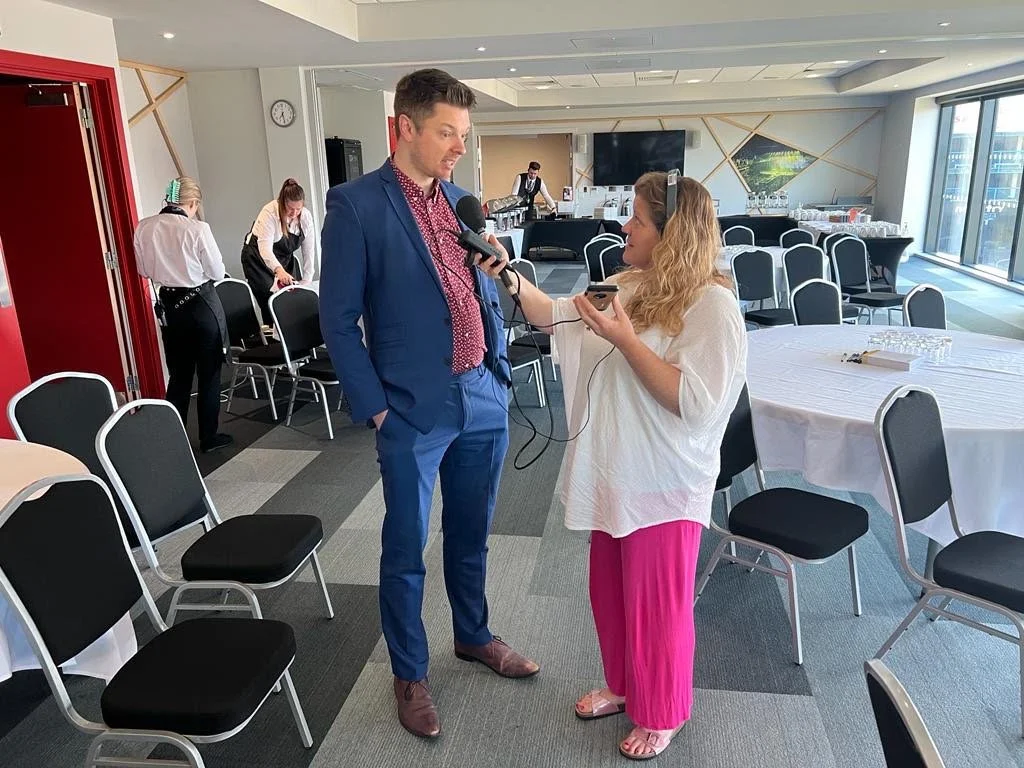 A man in a blue suit being interviewed by a woman holding a microphone and a smartphone in a conference room with empty chairs and tables, some set for an event.