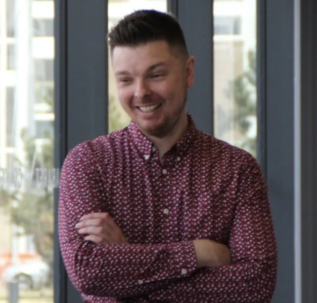 A man with short dark hair and a beard, smiling with arms crossed, looking down. He is wearing a red patterned button-up shirt and standing indoors near large glass windows with a view of buildings outside.