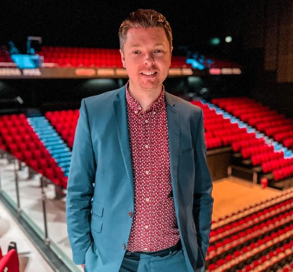 A man in a blue suit and patterned red shirt standing inside a theater with red seats and a stage in the background.