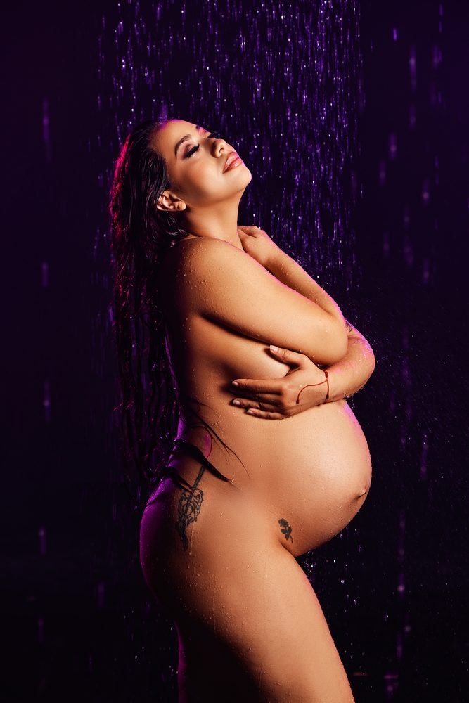 Maternity photography in Austin, Texas by Breeanna Rose Photography. Standing rain-room portrait, bare belly, arms crossed, purple light, head tilted back.