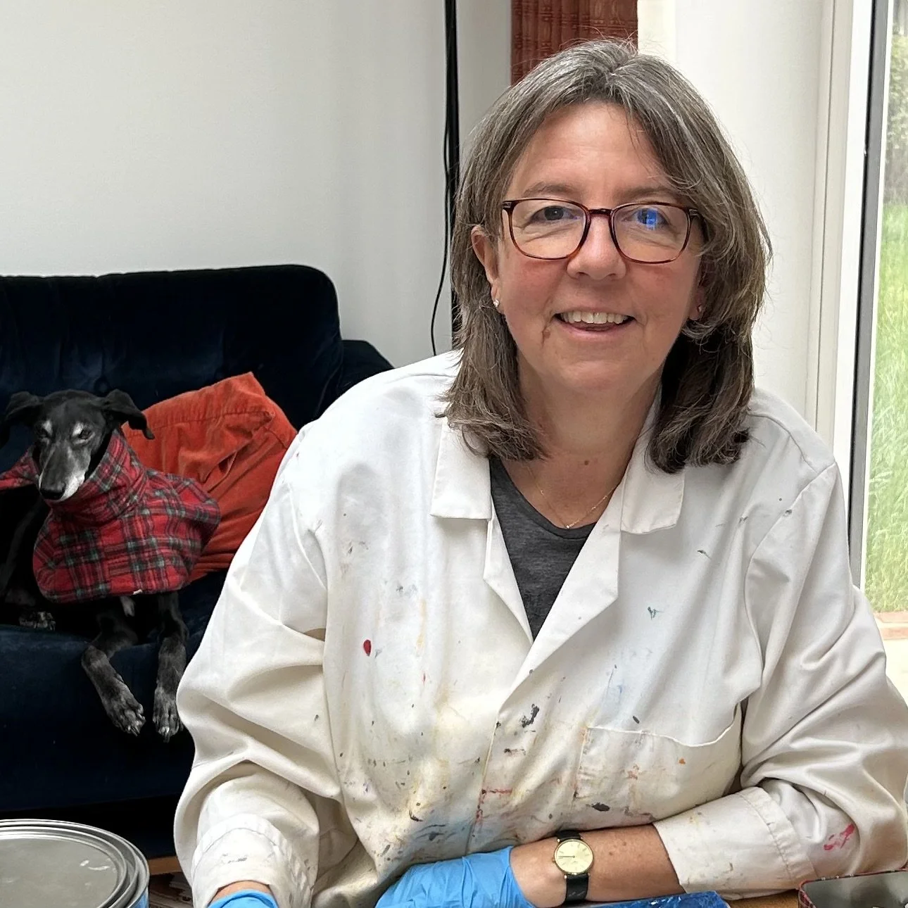 Kate Grimes smiling in her studio, wearing a paint-splattered white coat and blue gloves, with one of her dogs resting on the sofa behind her.