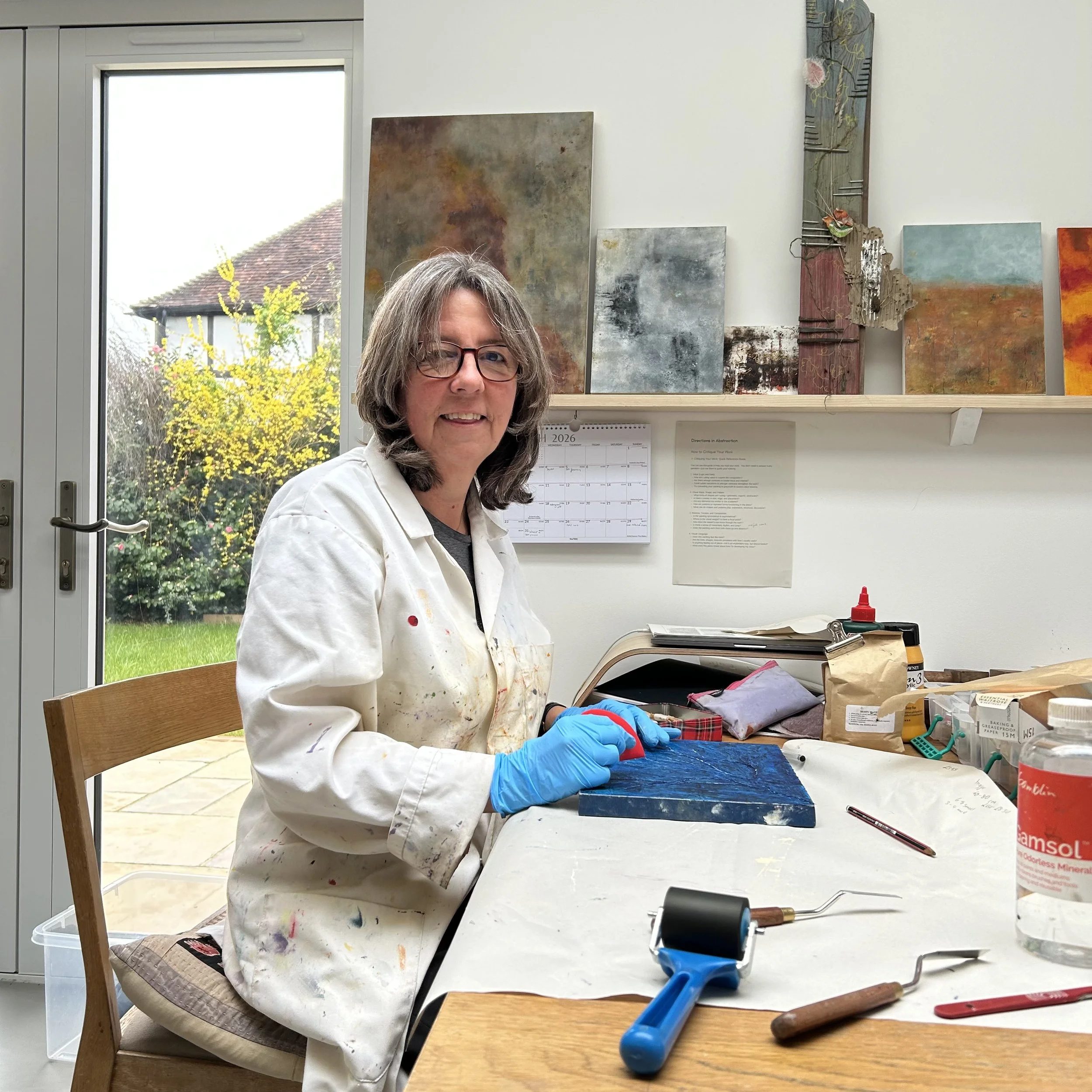 Kate Grimes smiling in her studio, wearing a white painting coat, with abstract paintings behind her.