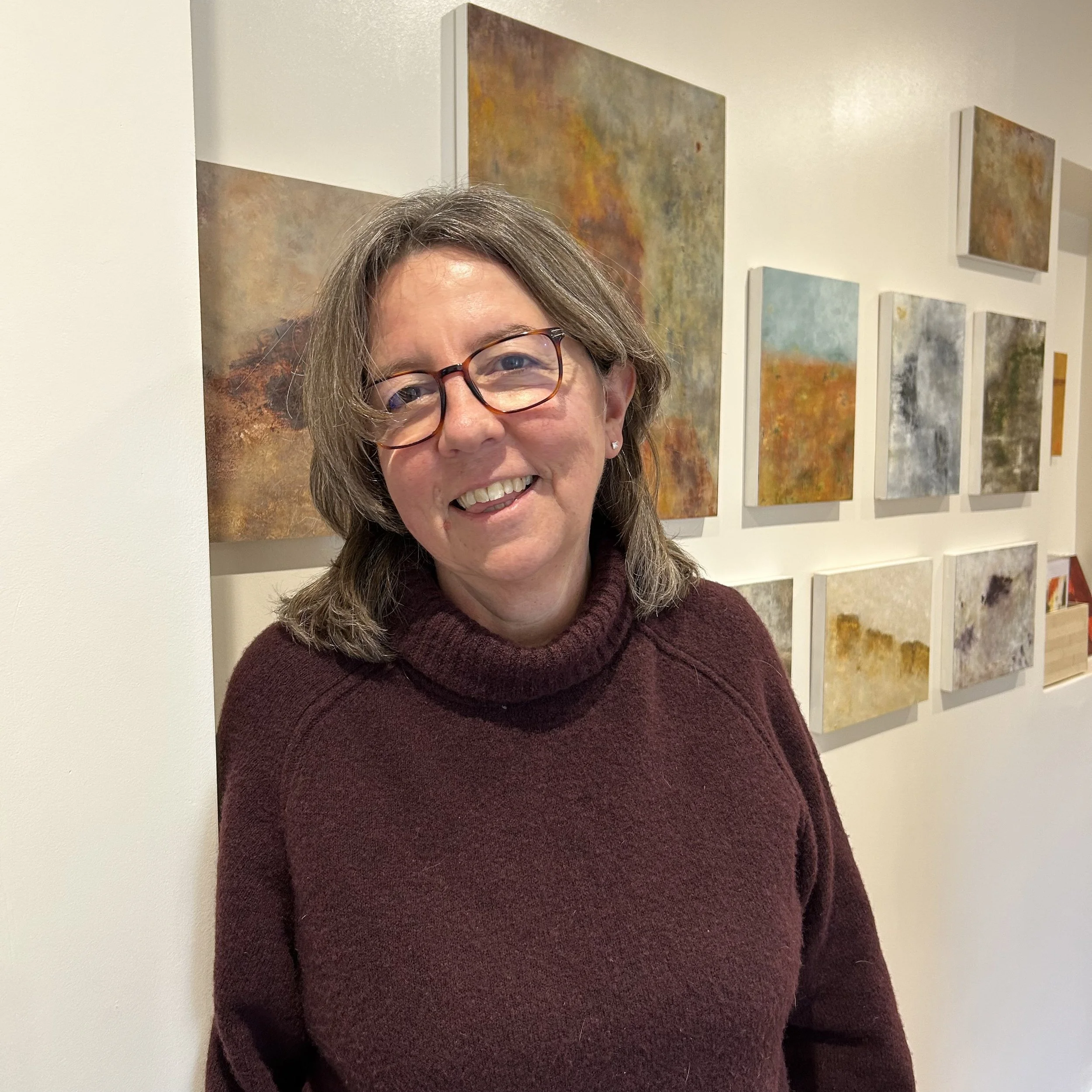 Kate Grimes smiling in front of abstract landscape paintings on a gallery wall.