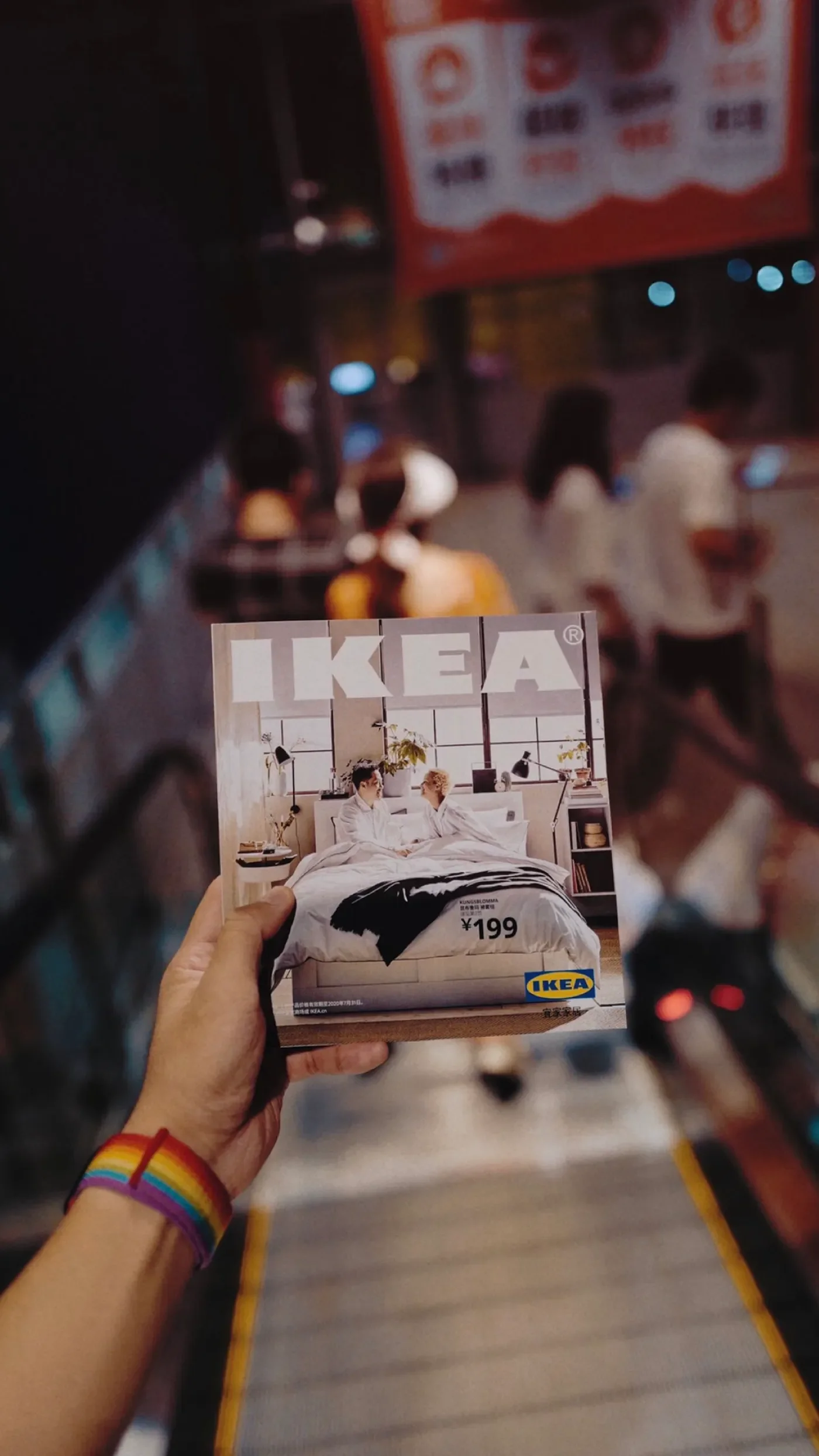 A person wearing rainbow-colored bracelets holds a magazine or catalog for IKEA featuring a bedroom setup, with people sitting on a bed and priced at ¥199. The background is blurred with people and illuminated signs.