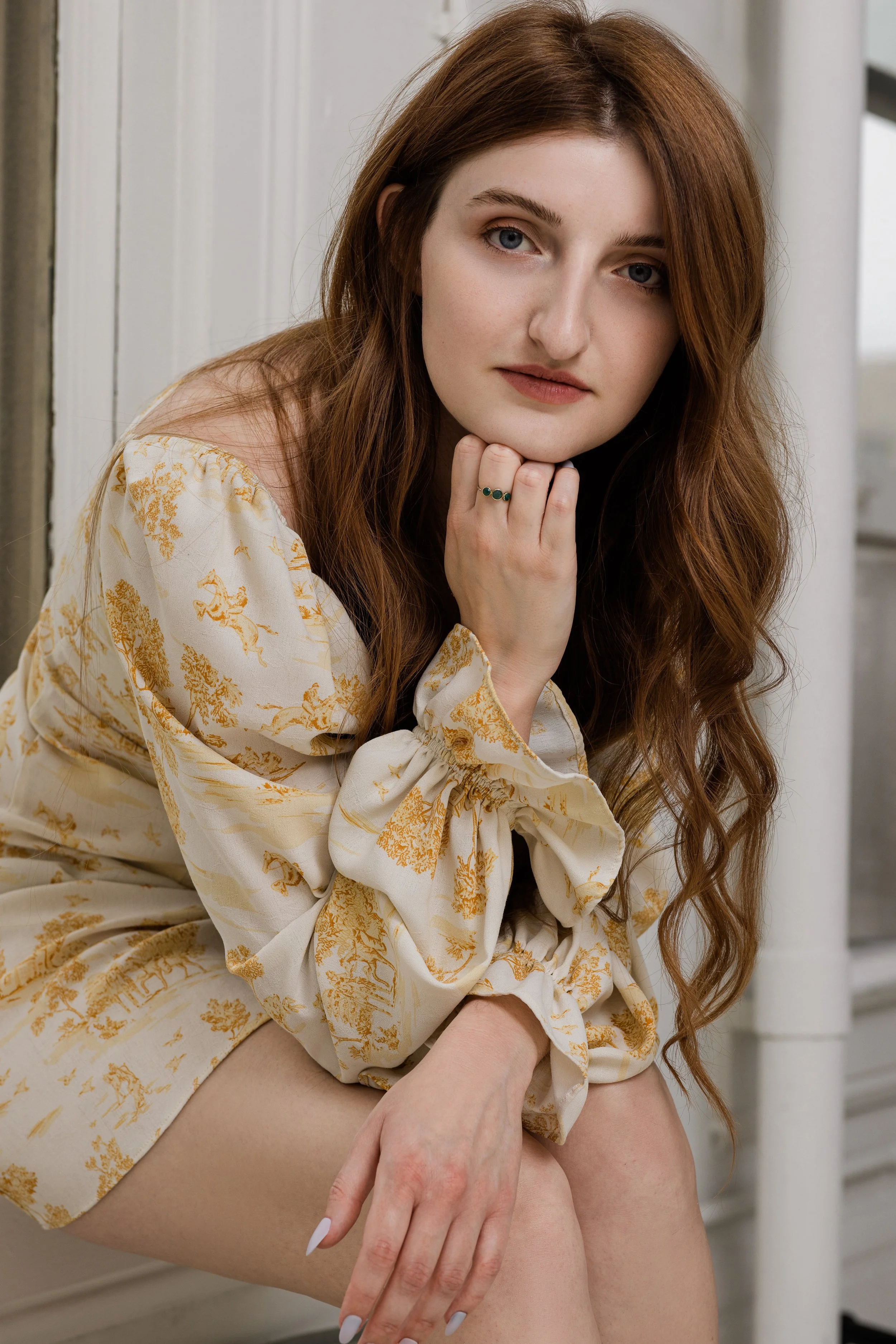 A woman with long red hair and light skin sitting near a window, wearing a cream and gold patterned dress with puffed sleeves, resting her chin on her hand.