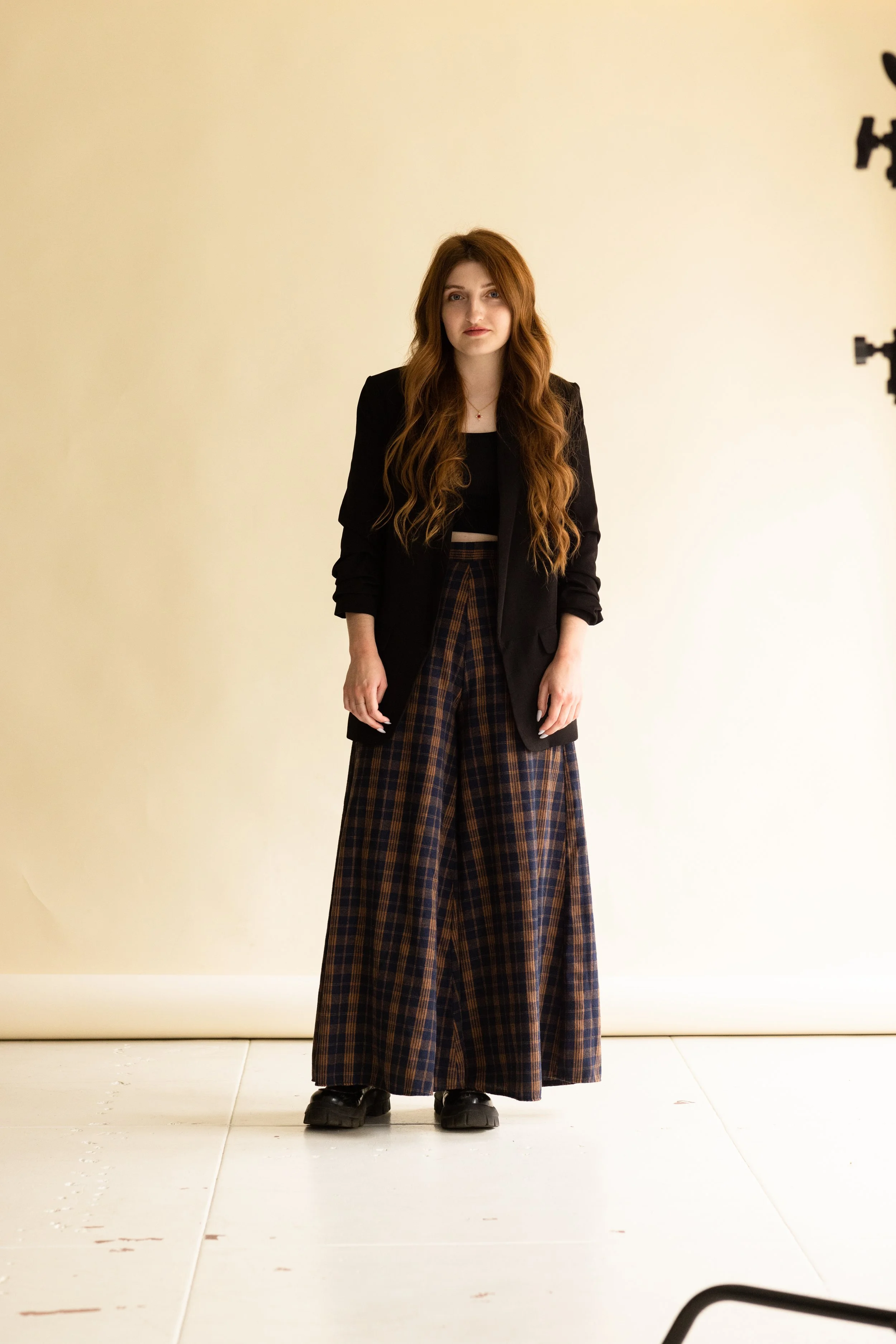 A young woman with long wavy red hair stands against a plain beige backdrop. She wears a black blazer, a black top, and wide-legged plaid pants with a brown and navy pattern. She also wears black shoes. She looks directly at the camera with a slight, neutral expression.