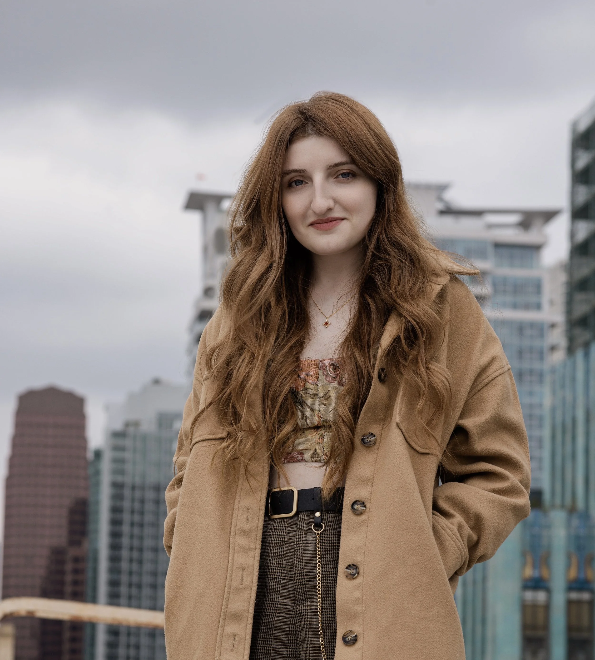 A woman with long red hair wearing a beige coat, patterned dress, and a belt stands outdoors against a cityscape with modern glass buildings on a cloudy day.