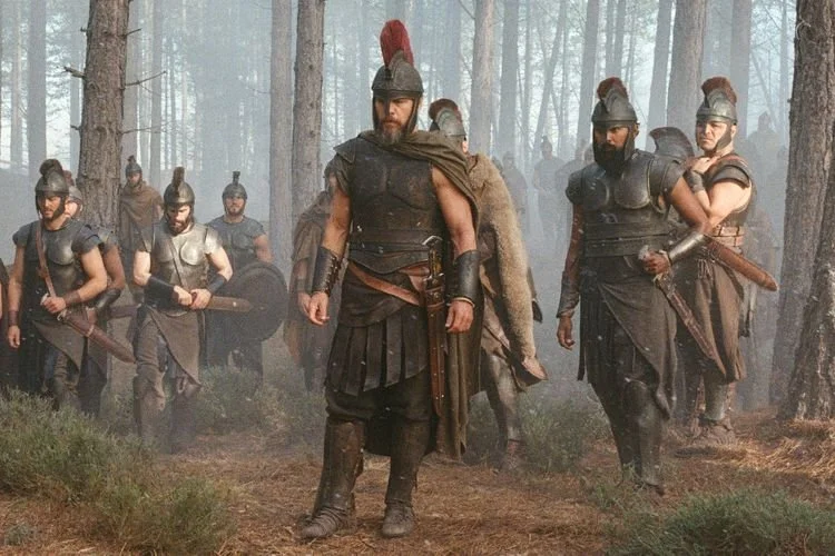 Group of Roman soldiers marching through a forest, wearing armor and helmets, carrying shields and weapons.