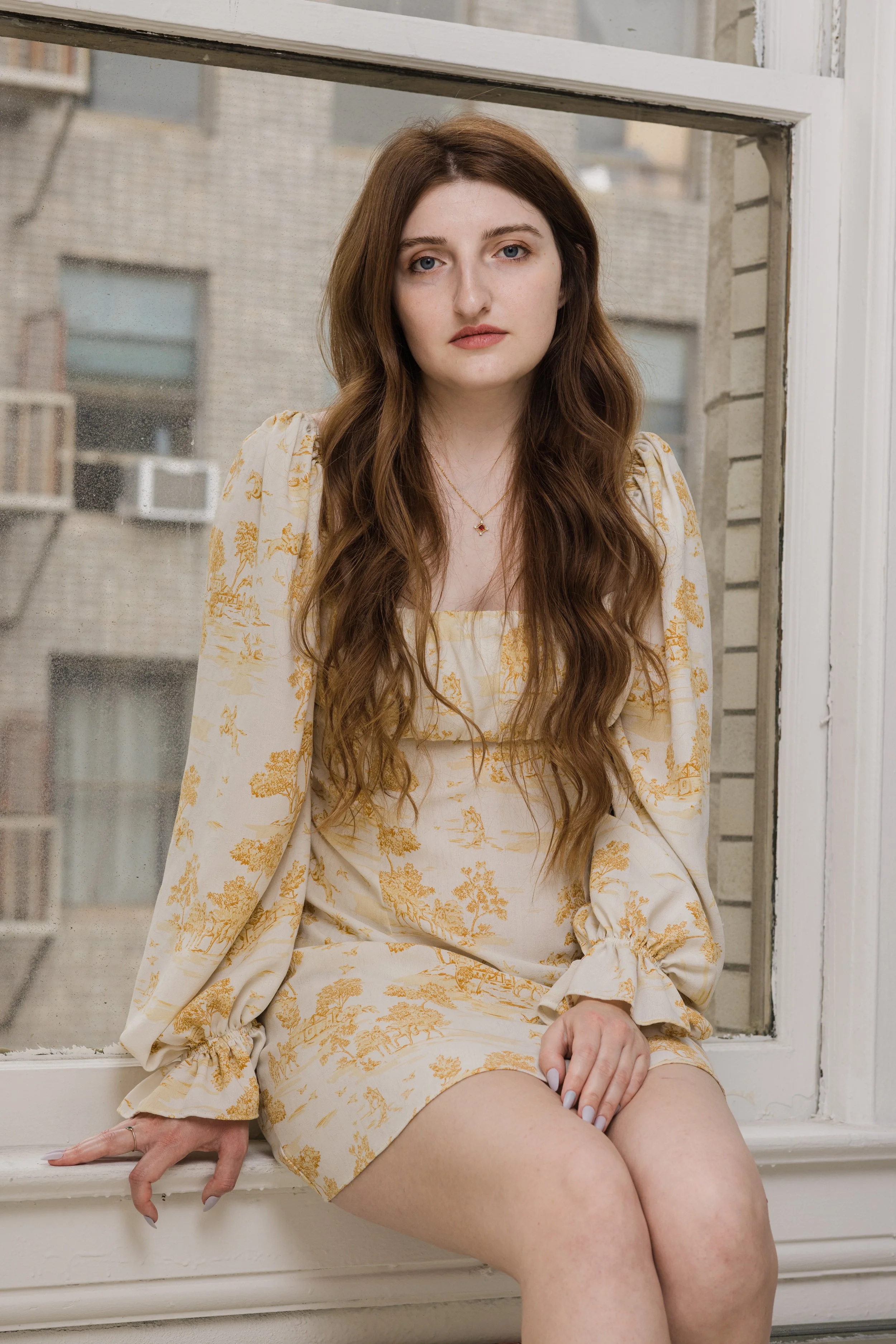 A young woman with long wavy brown hair and blue eyes sitting by a window, wearing a matching floral print dress and jacket in shades of cream and gold, with her hand resting on her knee and the other on the window ledge.