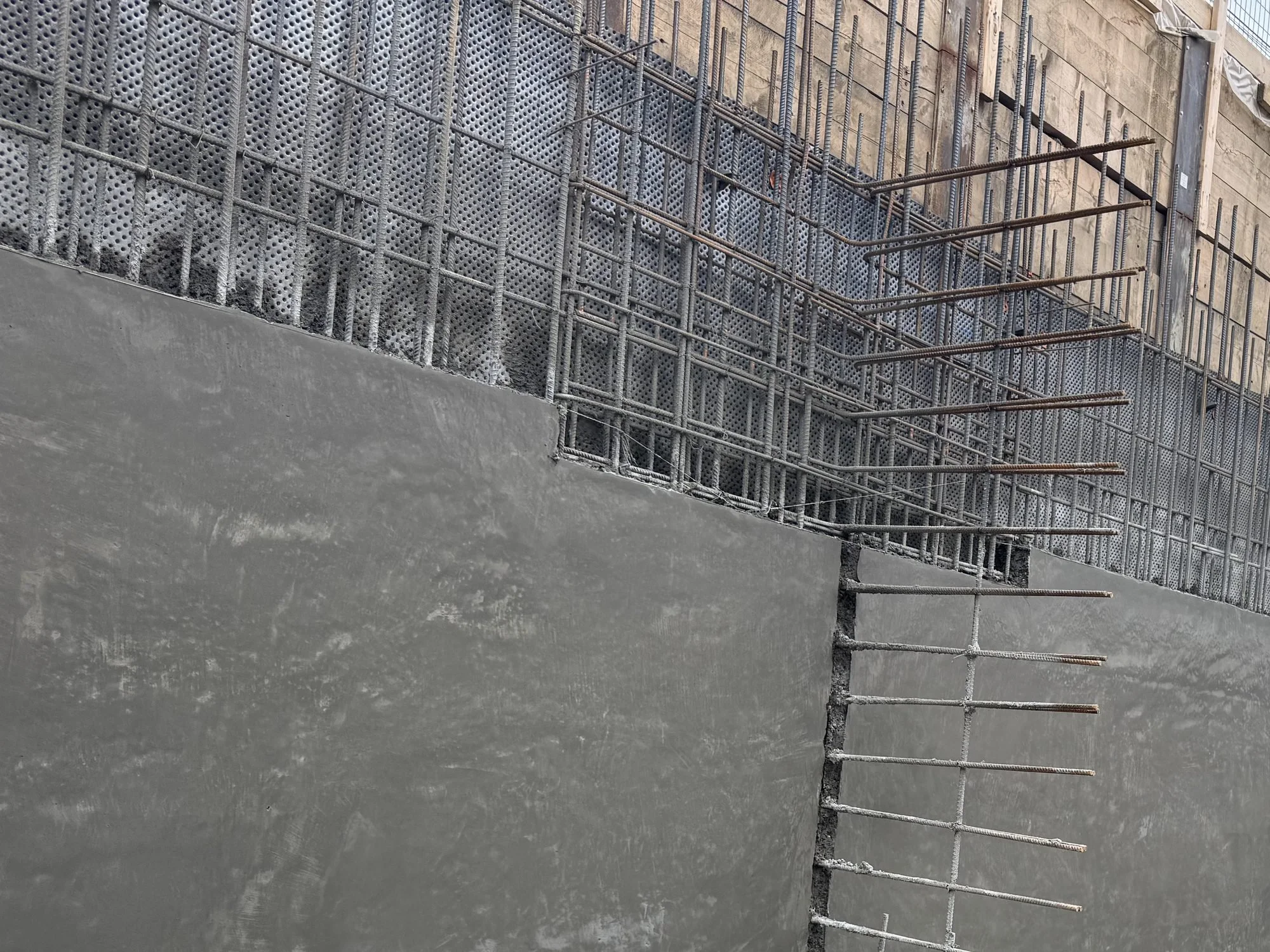 Rebar mat tied over dimpled drainage board against timber shoring, above a finished shotcrete wall, with horizontal dowels at a wall corner