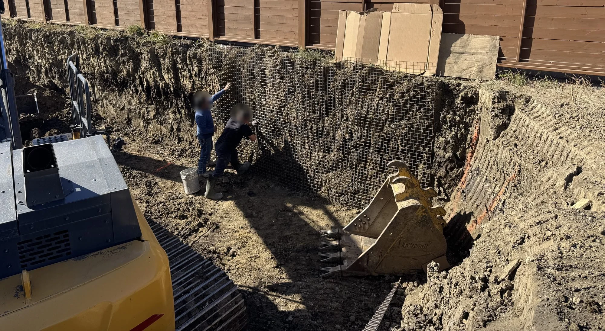 Crew fixing welded wire mesh to a soil cut along a wooden fence in a residential basement excavation, excavator bucket in the foreground