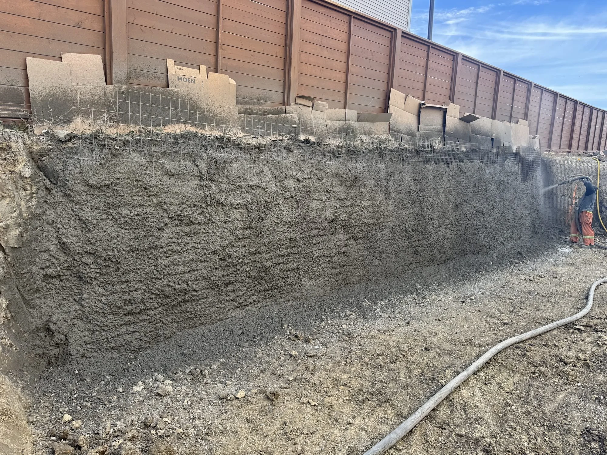 Nozzleman spraying wet-mix shotcrete onto a mesh-reinforced excavation face along a fence line, seen from behind