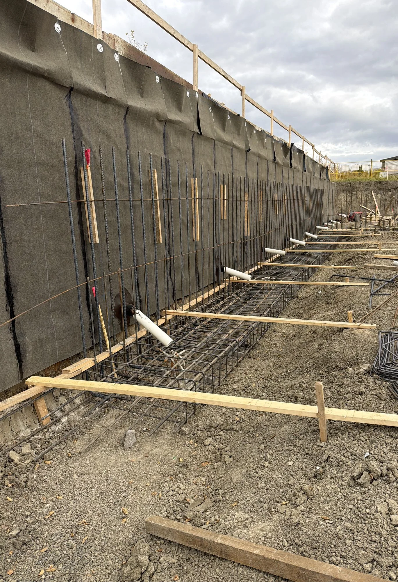 Rebar mats tied over black membrane against shoring, with pipe sleeves and footing steel at the base, before shotcrete