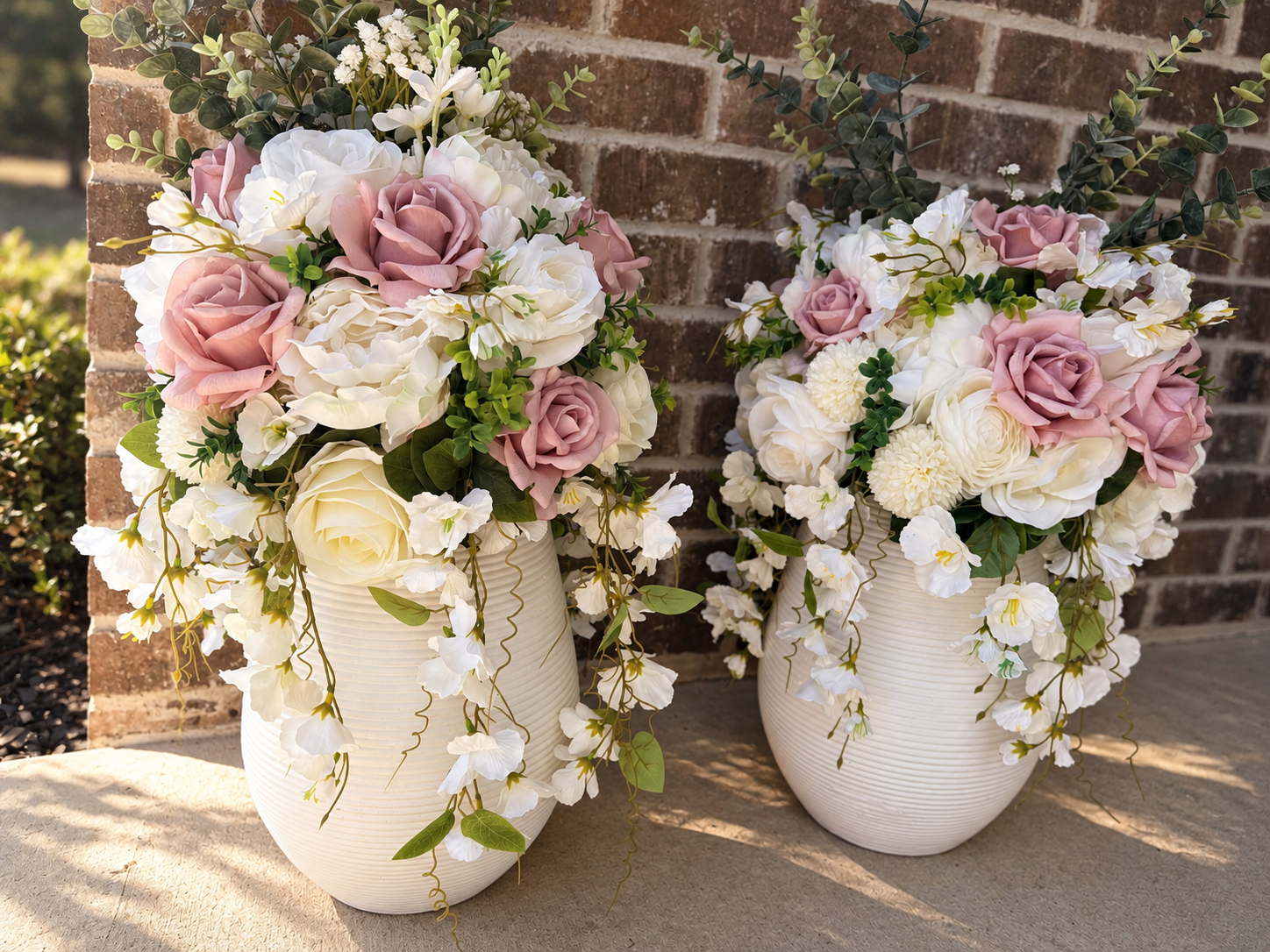 Two white vases filled with pink and white roses, white orchids, and green foliage, placed outdoors on a stone surface against a brick wall.