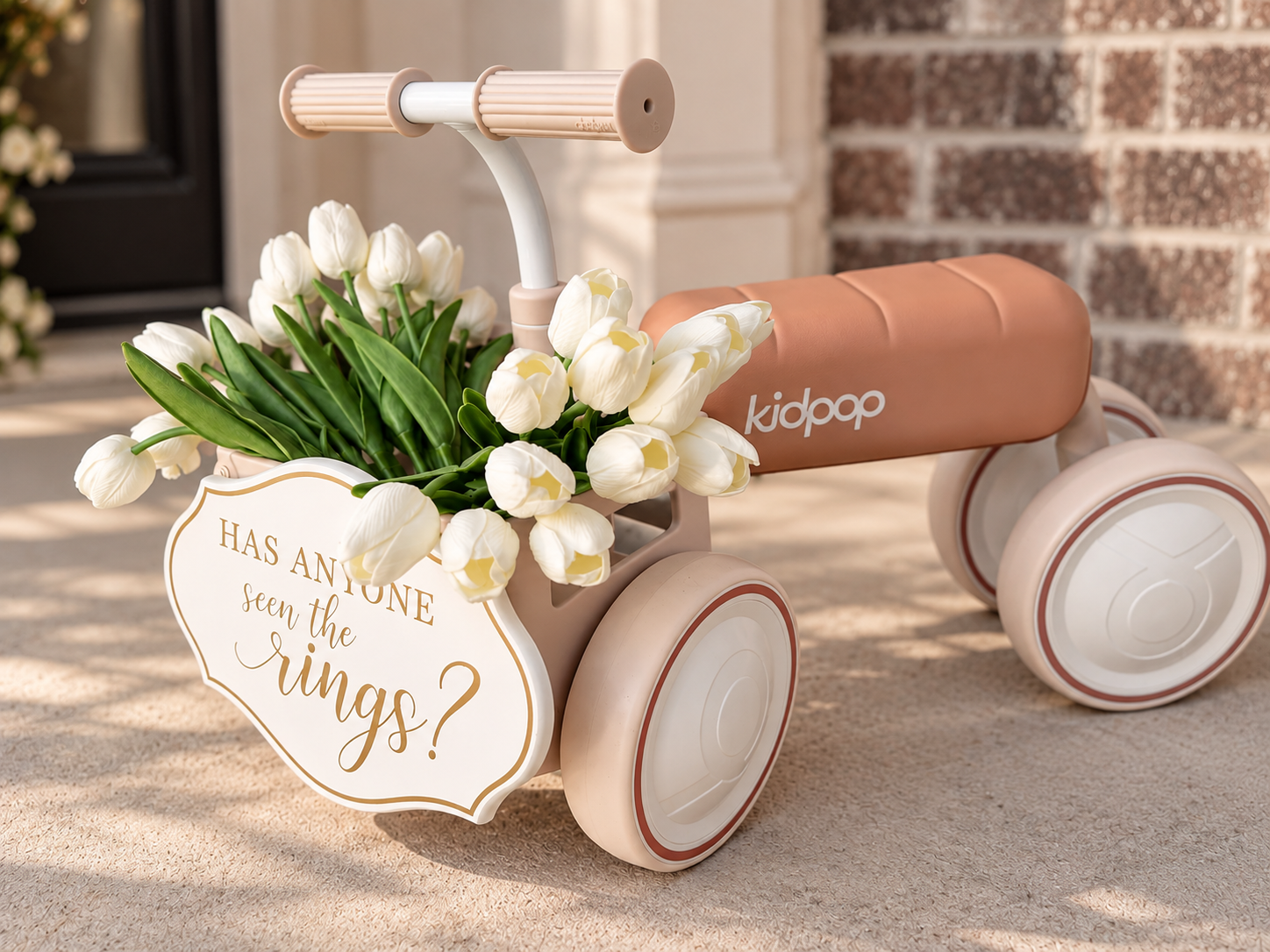 An ivory children's ride-on toy cart with white tulips and green leaves, a sign saying 'Has anyone seen the wings?', and a brown cushioned seat branded 'kidpop', placed outdoors on a sidewalk.