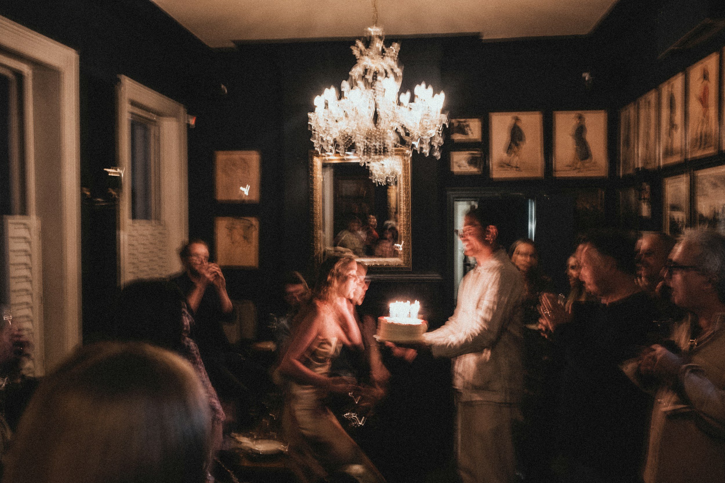 People gathered in a dimly lit room for a celebration, with a woman and a man holding a cake with candles, a chandelier hanging from the ceiling, and framed pictures on the dark wall.