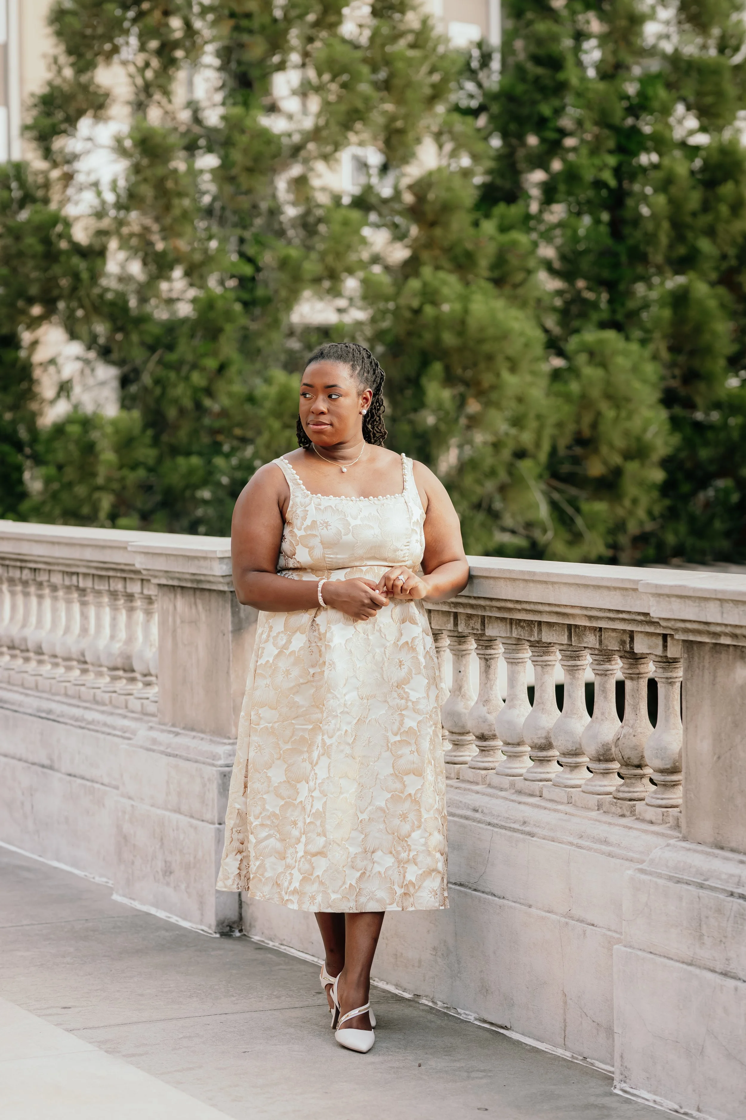 A woman in a cream-colored floral dress with spaghetti straps standing on a stone balcony with a stone railing, overlooking trees and buildings in the background.