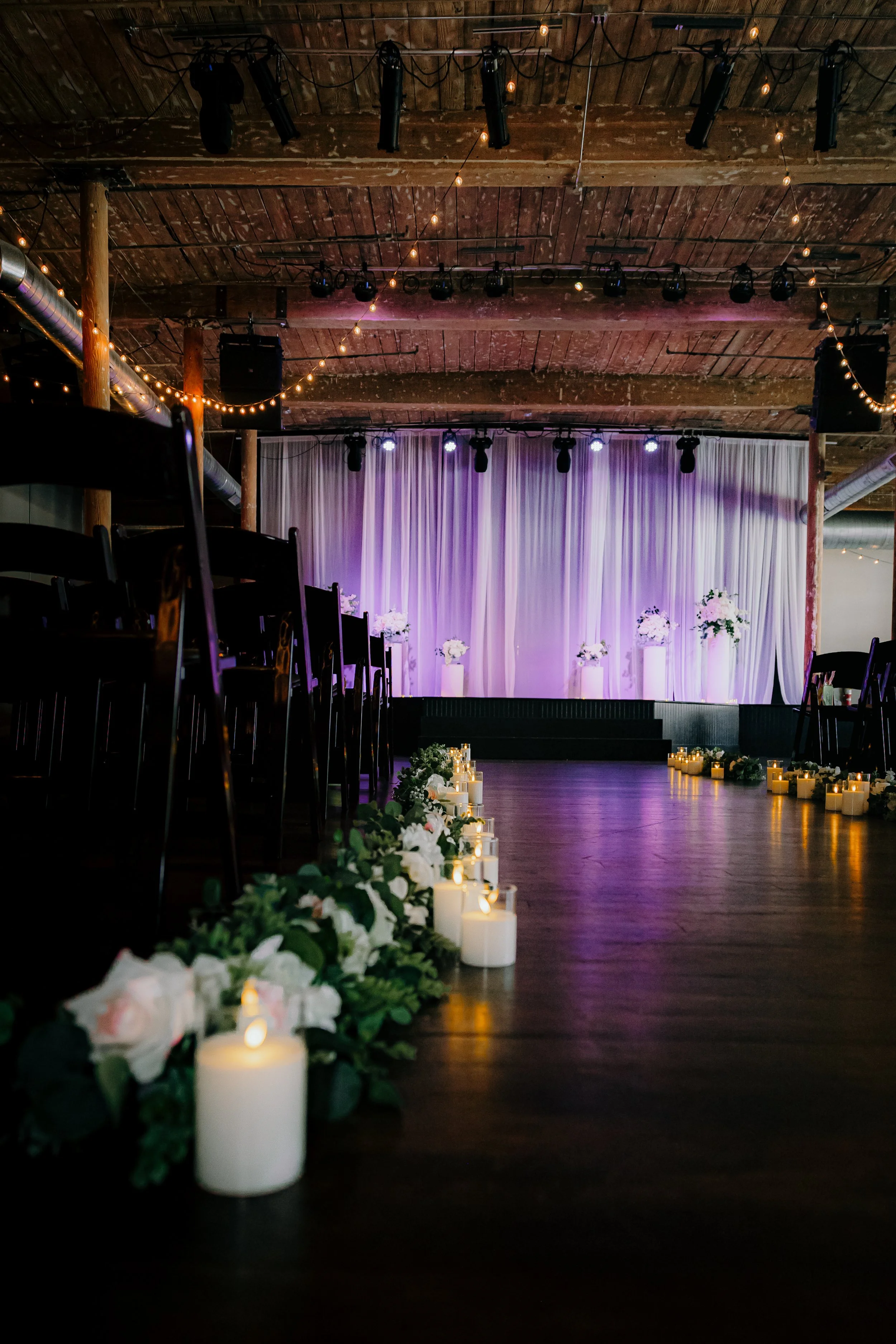 Empty event venue with candlelit floral arrangements along the aisle, a purple-lit stage with white curtains, wooden ceiling with hanging string lights, and chairs arranged for a wedding or celebration.