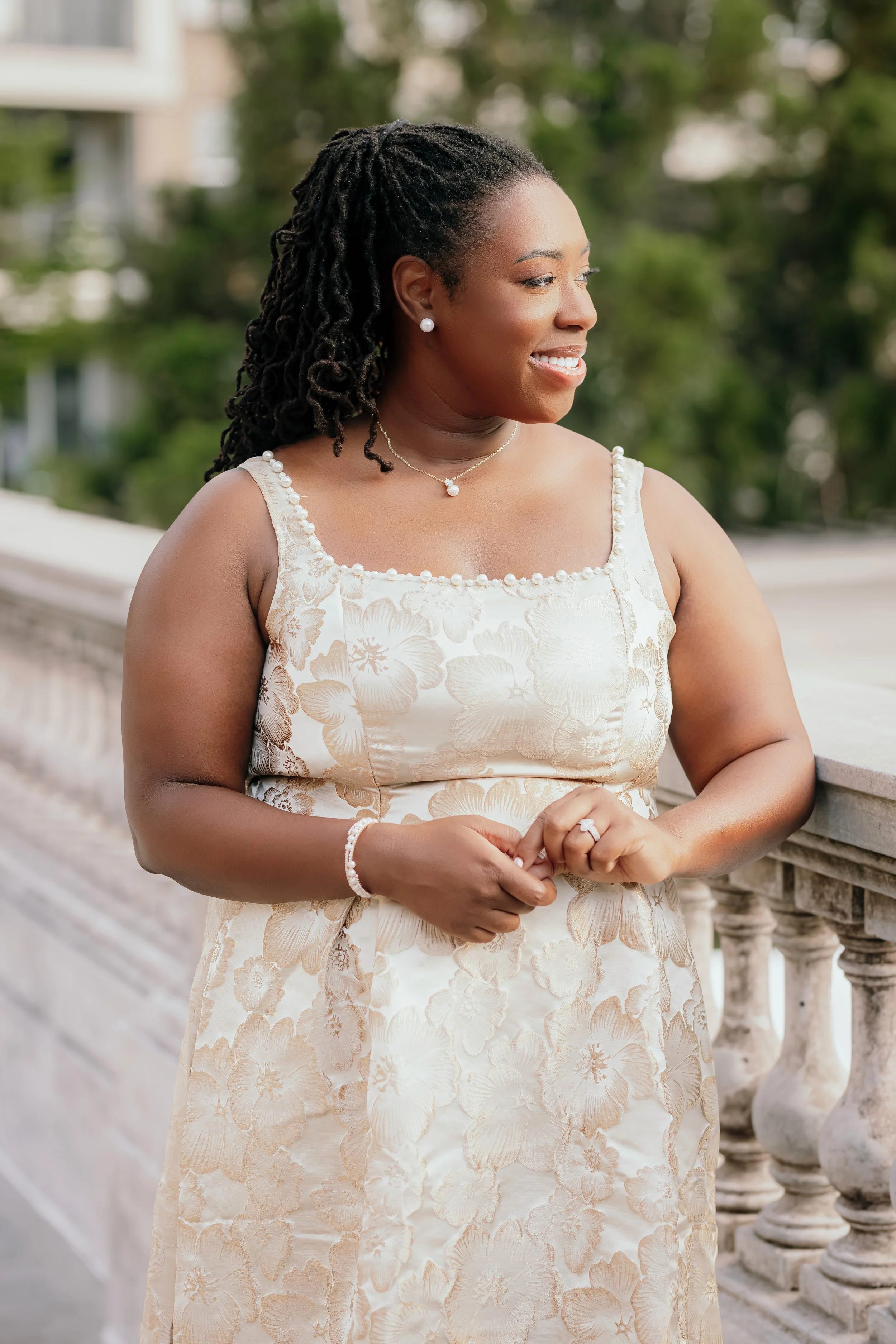 A woman with dark, curly hair wearing pearl earrings and a matching pearl necklace, dressed in a cream-colored floral dress, standing outdoors on a stone bridge with greenery in the background, smiling and looking to her right.