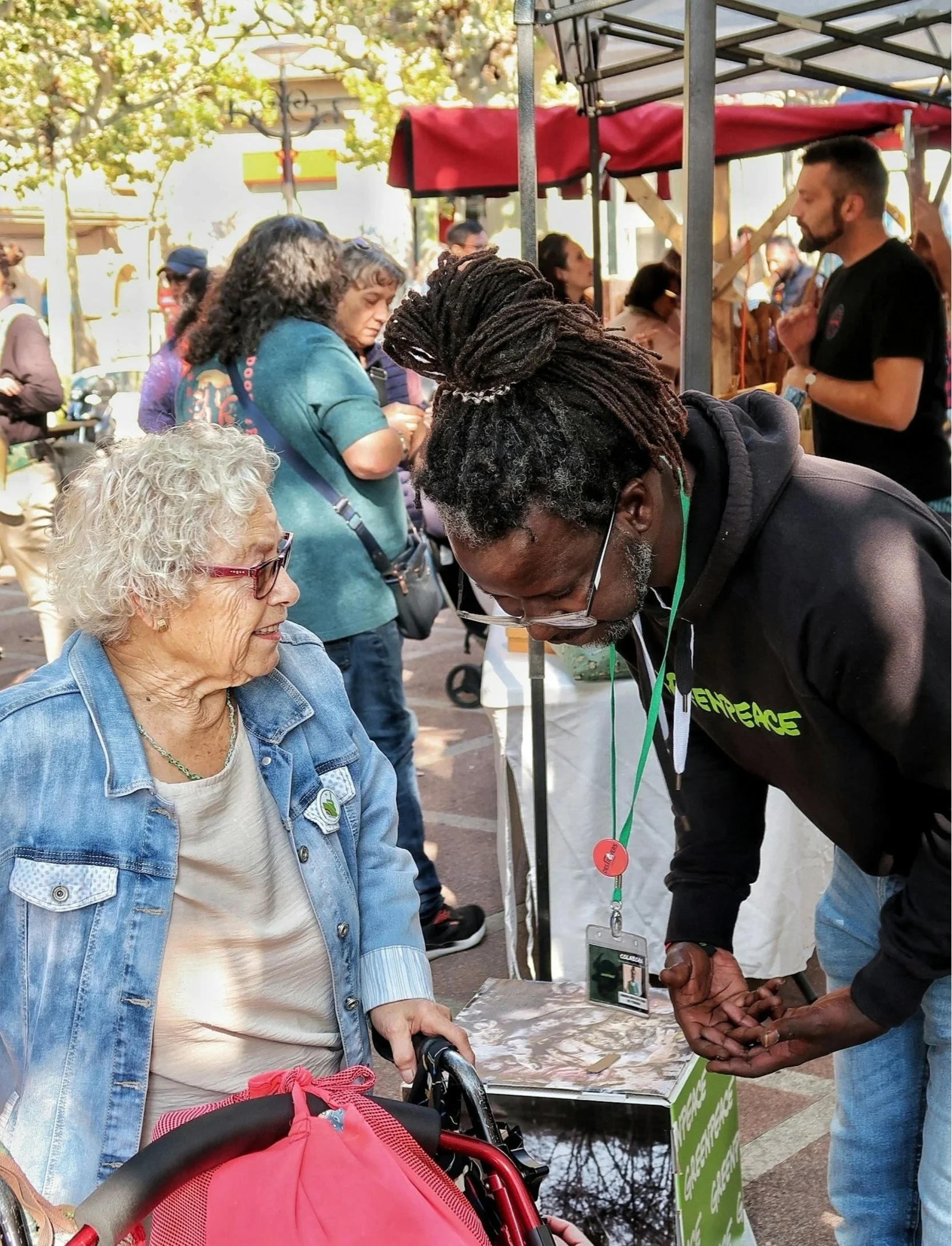 People at an outdoor event, including a woman in a denim jacket sitting in a wheelchair, and a man wearing a black hoodie engaging in conversation. There are multiple individuals in the background, with some looking at their phones. A red stroller is in the foreground, and a tent or booth with a red cover is visible. The scene takes place during daytime with trees and buildings in the background.