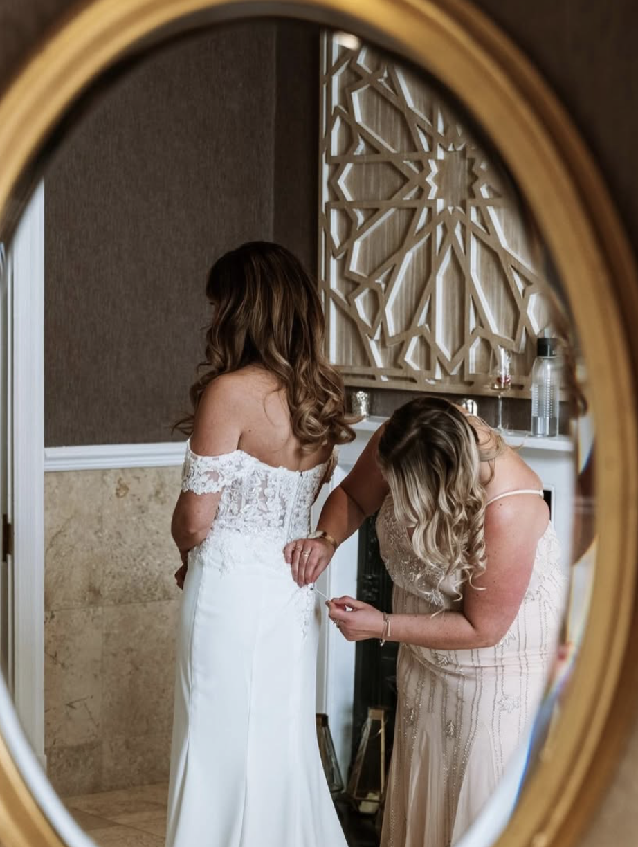 Woman in a strapless wedding gown getting assistance from another woman in a cream-colored dress, reflected in a mirror with an ornate wooden frame.