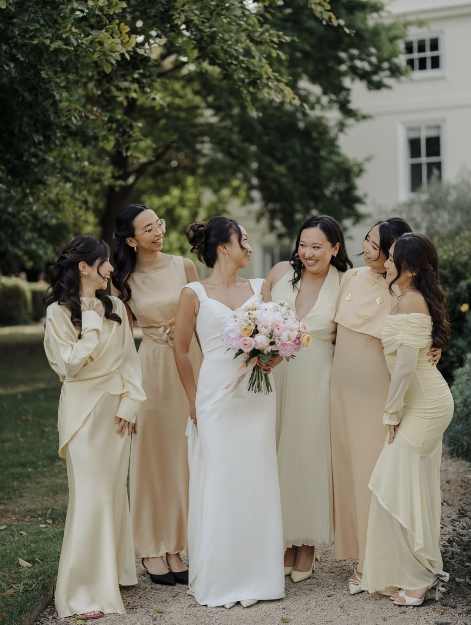 A group of six women, including a bride in a white wedding dress holding a bouquet, standing outdoors in a garden with trees and a white building in the background, smiling and interacting warmly.