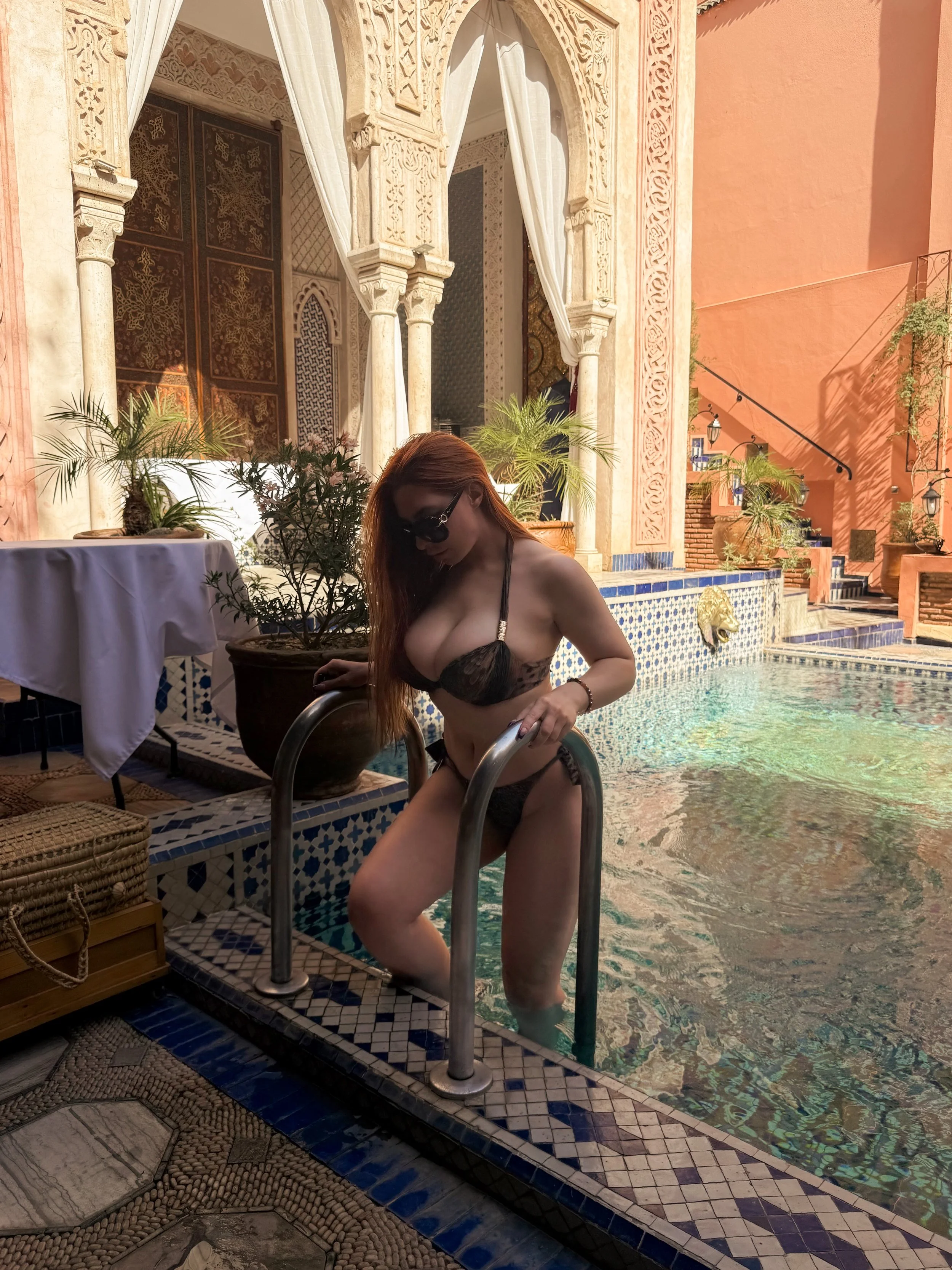 A woman with red hair wearing sunglasses and black lingerie descending a pool ladder in a luxurious Moroccan-style indoor pool area, decorated with ornate arches, plants, and intricate tilework.