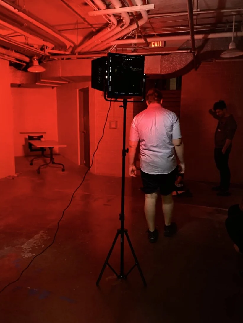 Indoor scene with red lighting, a large video monitor on a stand, a man in a white shirt and black shorts, and other people in the background, possibly preparing for a video shoot or event.