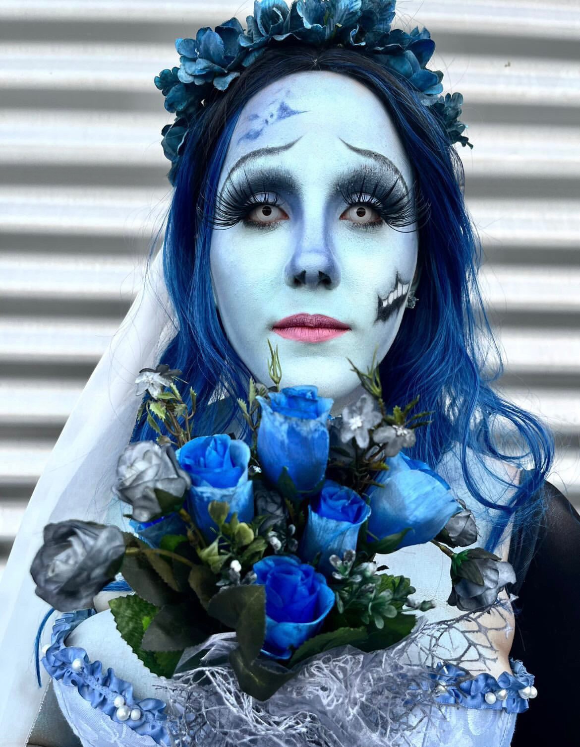A woman dressed in blue-themed makeup and costume holds a bouquet of blue and gray roses. She has long blue hair, elaborate eye makeup, and a floral headpiece, with a skeletal face design on one side of her face.