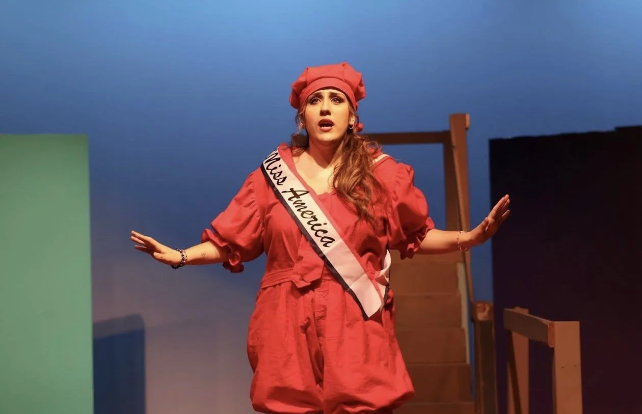 A woman performing on stage, dressed in a red costume with a sash that reads 'Miss America,' wearing a red nurse or captain's hat, with colorful lighting and a simple stage set.