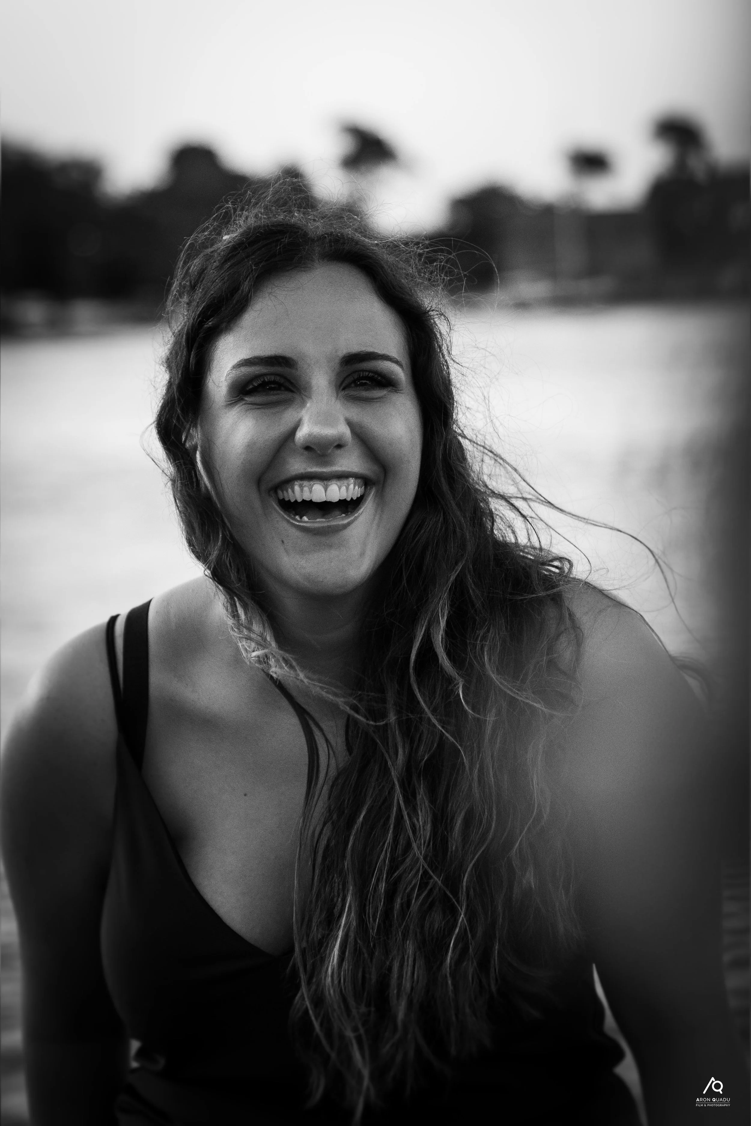 A woman with long wavy hair smiling broadly, wearing a sleeveless top, outdoors near a body of water, with blurred trees and structures in the background.