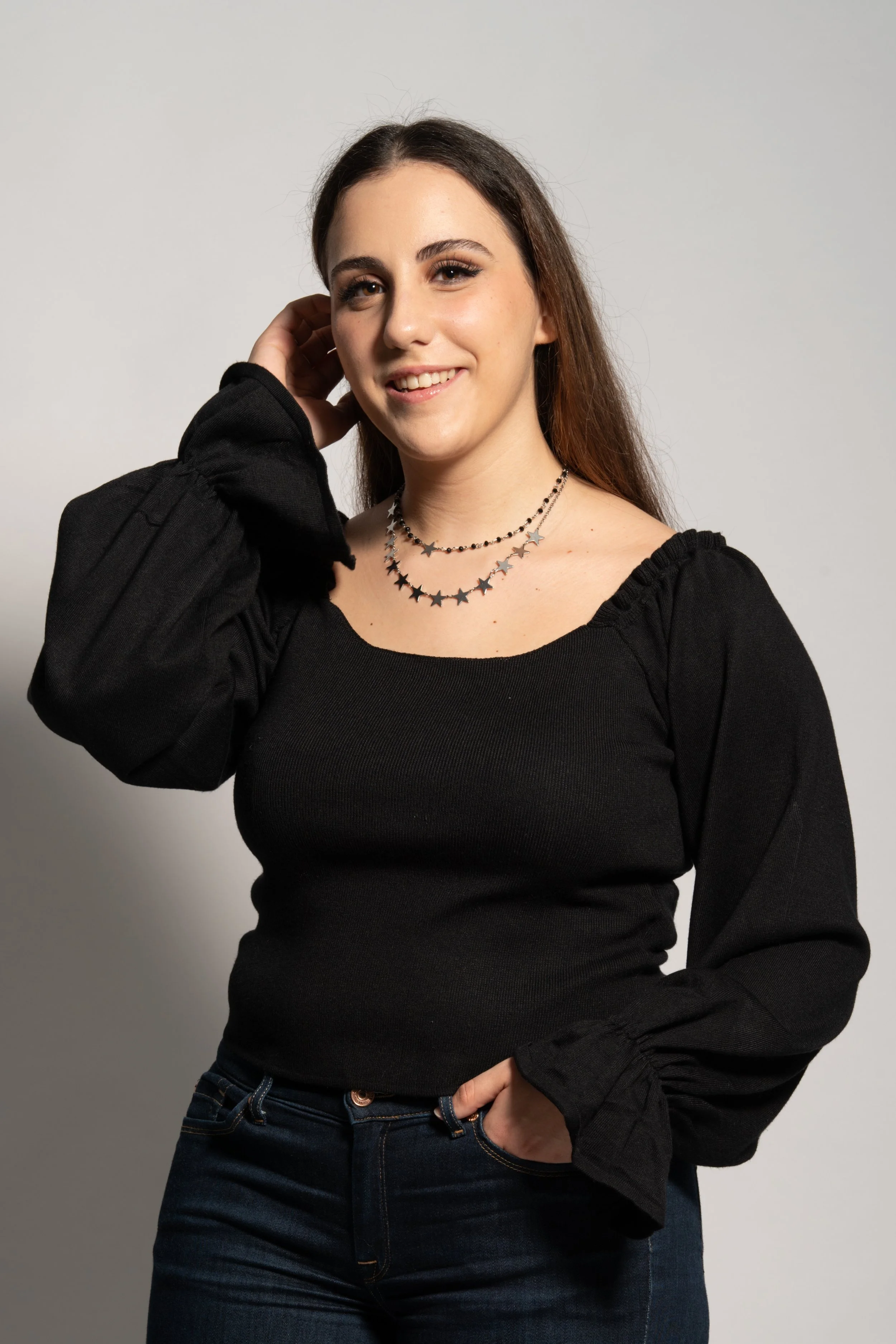 A young woman with long brown hair wearing a black long-sleeve top with puffed sleeves and a layered star necklace, standing against a plain gray background. She is smiling with one hand touching her head.