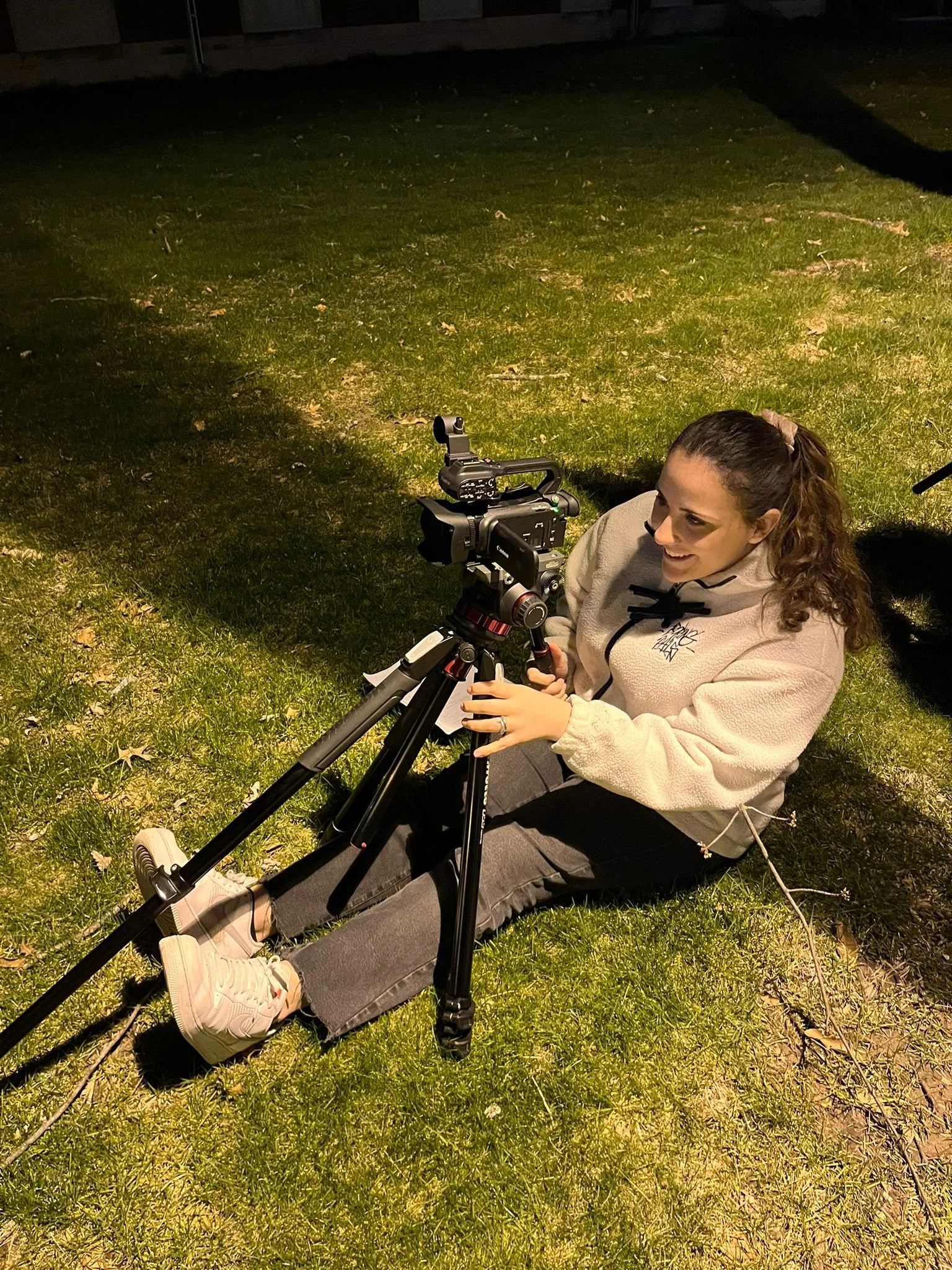 A woman sitting on the grass at night operating a professional camera on a tripod.