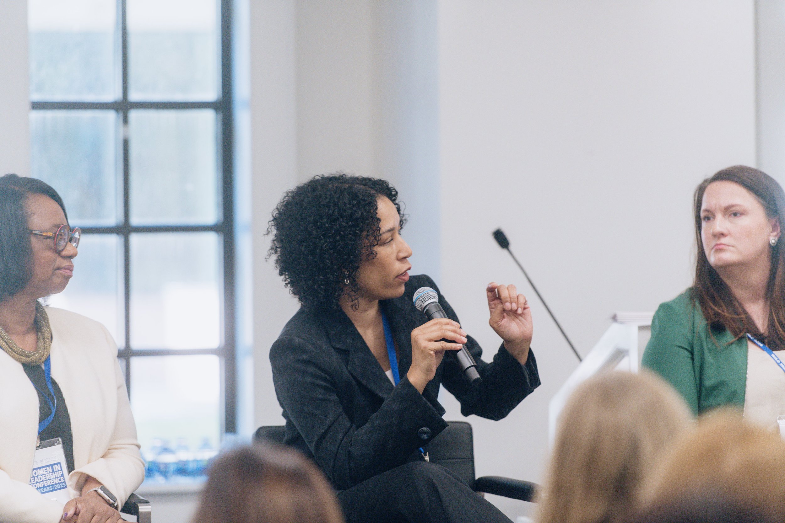 A woman with curly black hair speaking into a microphone during a conference or panel discussion, sitting among other women in a bright room with large windows.