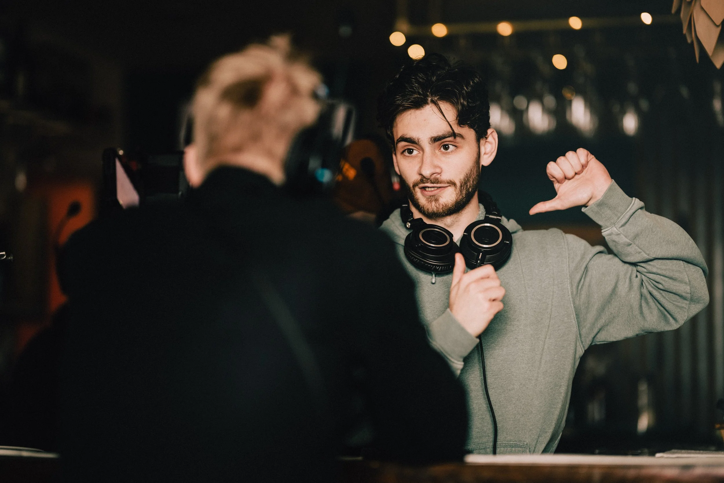 A young man with dark hair and a beard wearing a gray hoodie, headphones around his neck, and gesturing with his hand, appears to be talking to another person with light hair, who is mostly out of focus. The scene is indoors with blurry string lights in the background.